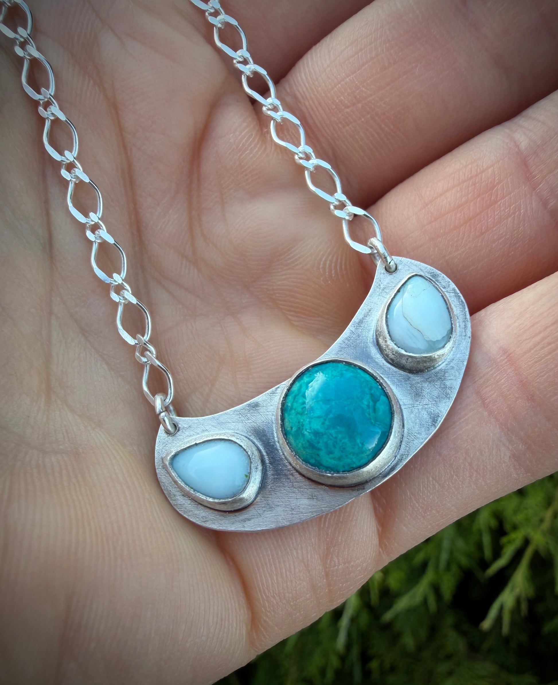 A close-up of the handcrafted sterling silver Marisol Pendant held in a hand. The tiered pendant features a central, circular teal Chrysocolla stone flanked by two smaller, sky-blue Larimar stones. The metal has a weathered, matte texture that contrasts with the polished silver bezels and the delicate, elongated link chain.