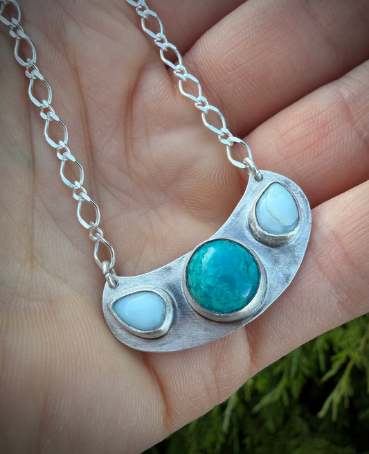 A close-up of the handcrafted sterling silver Marisol Pendant held in a hand. The tiered pendant features a central, circular teal Chrysocolla stone flanked by two smaller, sky-blue Larimar stones. The metal has a weathered, matte texture that contrasts with the polished silver bezels and the delicate, elongated link chain.