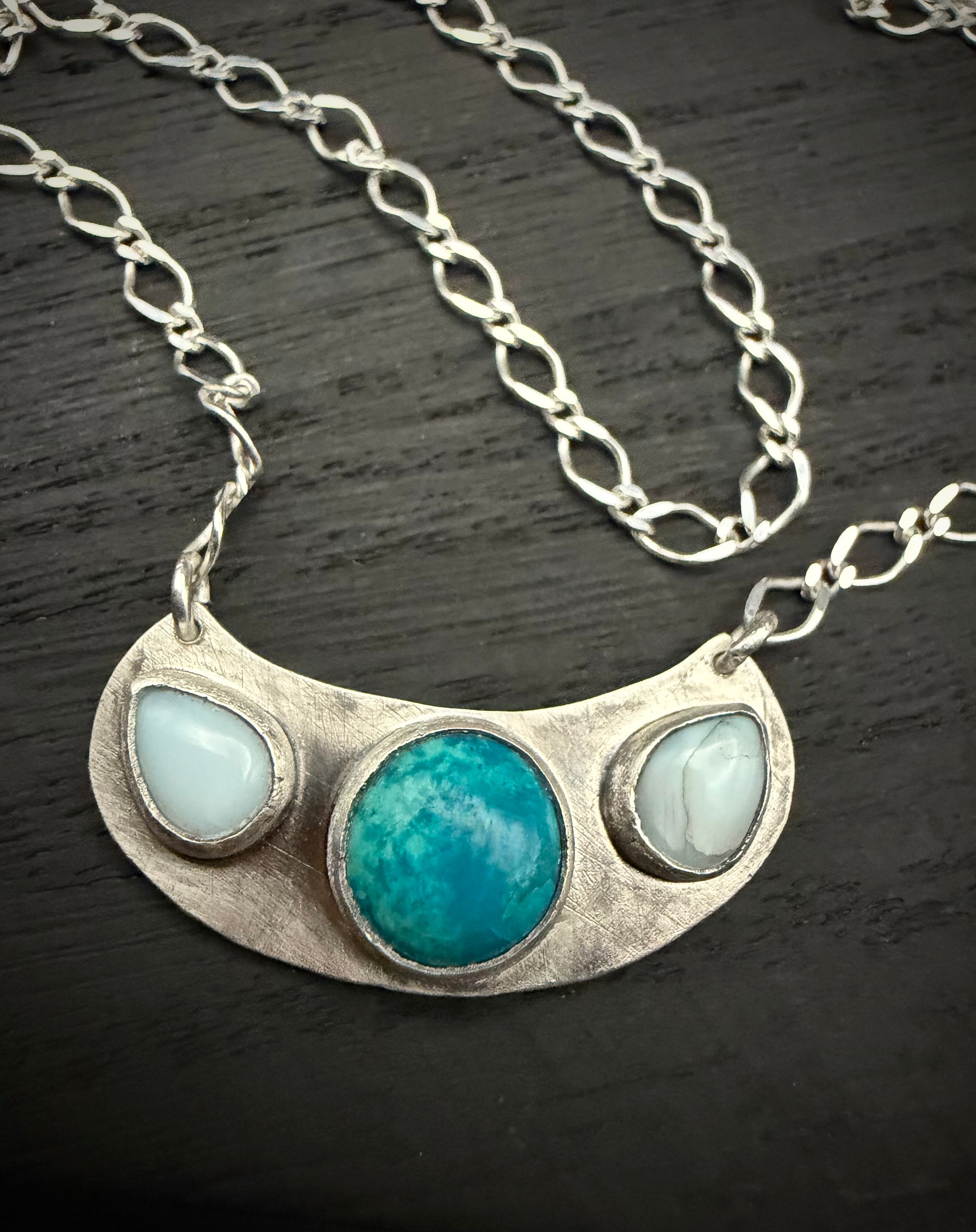 A close-up of the handcrafted sterling silver Marisol Pendant resting on a dark, weathered wooden surface. The image showcases the large, circular teal Chrysocolla stone in the center, flanked by two smaller, pale-blue Larimar stones on a crescent-shaped silver base. The long-link silver chain is visible, draped across the dark background to highlight the pendant's architectural silhouette and matte finish.