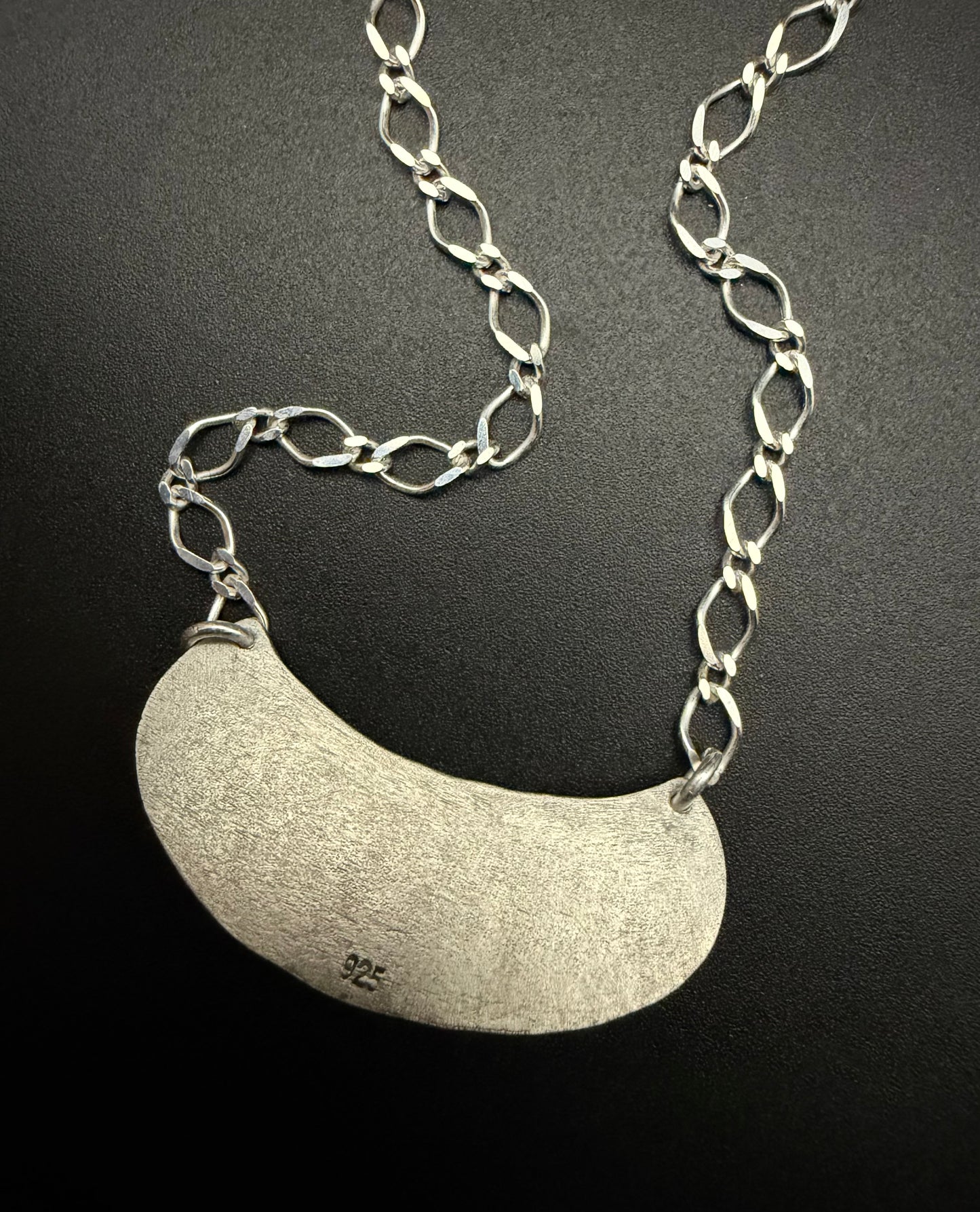 The reverse side of the handcrafted sterling silver Marisol Pendant against a dark, matte background. The image highlights the weathered, textured silver crescent. The long-link sterling silver chain is draped above the pendant, showcasing the sturdy construction and the matte, architectural finish of the metal.