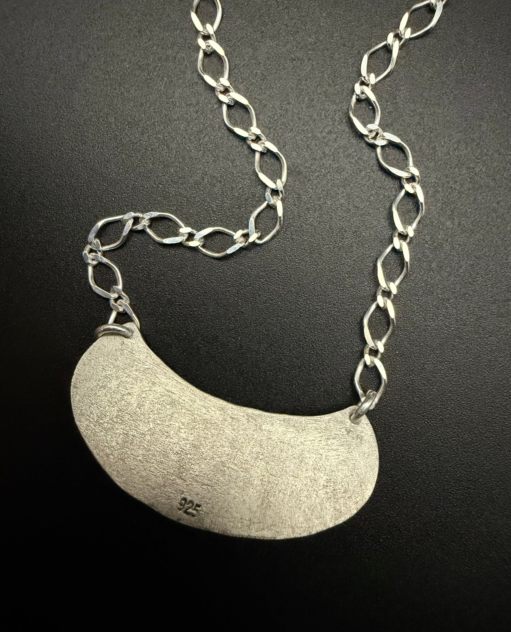The reverse side of the handcrafted sterling silver Marisol Pendant against a dark, matte background. The image highlights the weathered, textured silver crescent. The long-link sterling silver chain is draped above the pendant, showcasing the sturdy construction and the matte, architectural finish of the metal.