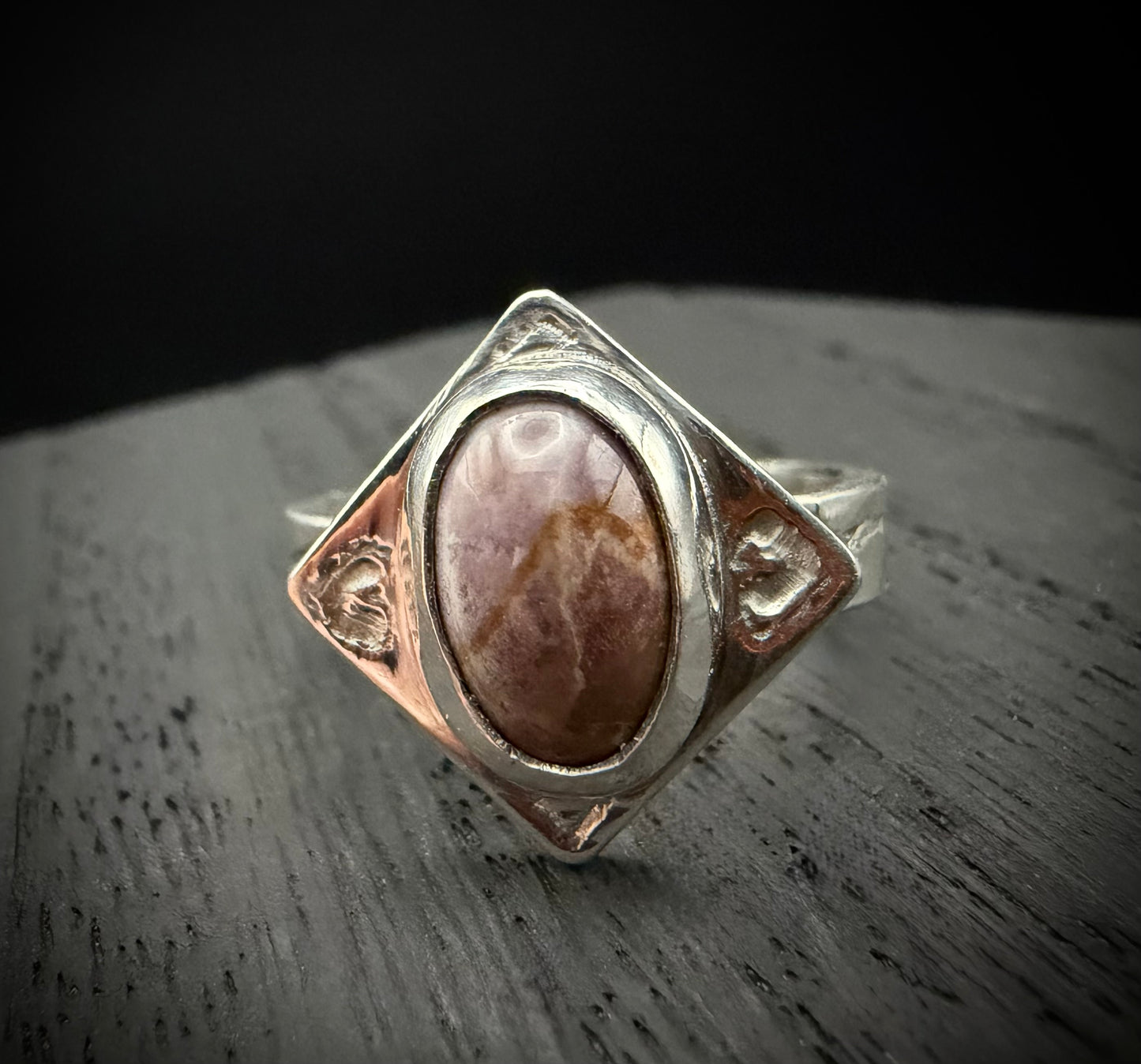 A front-facing view of the handcrafted sterling silver Tephra Ring on a dark, textured wood surface. The image emphasizes the oval brown agate stone centered within a diamond-shaped plate, showcasing the engraved heart motifs and the contrast between the polished silver bezel and the raw, molten metal texture.