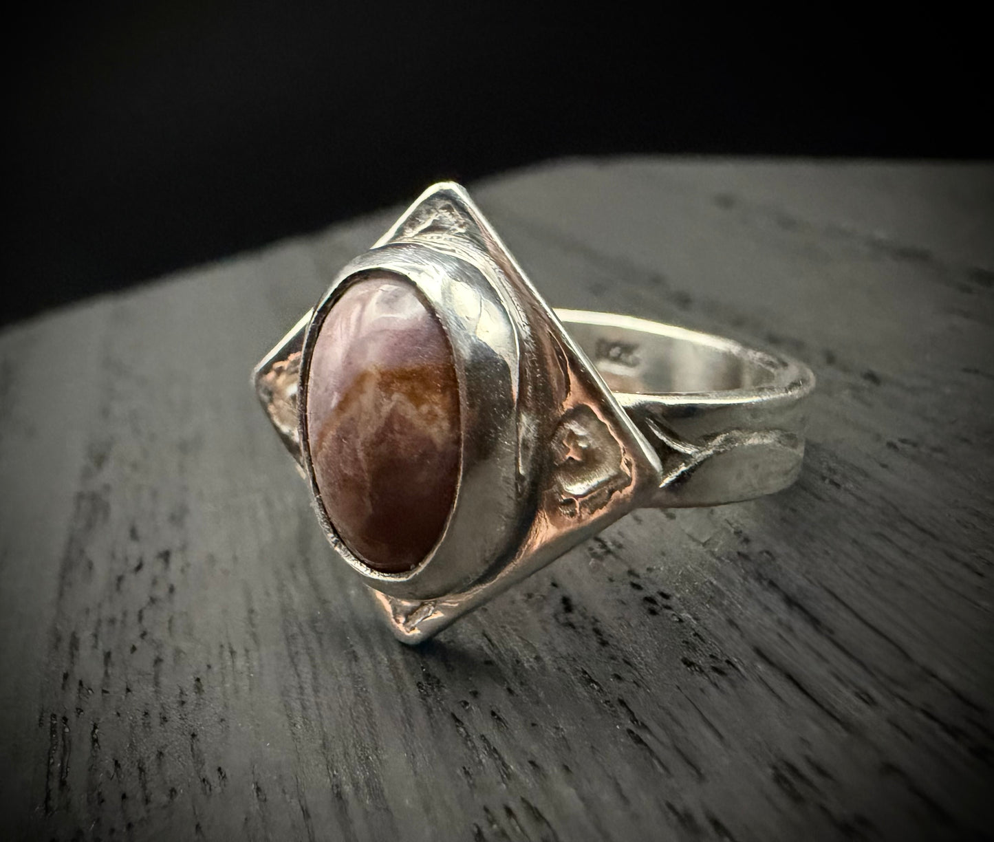 A close-up of the handcrafted sterling silver Tephra Ring resting on a dark, weathered wood surface. The ring features a diamond-shaped plate with molten textures and an earth-toned agate stone, highlighting the contrast between the polished silver edges and the deep brown of the gemstone.