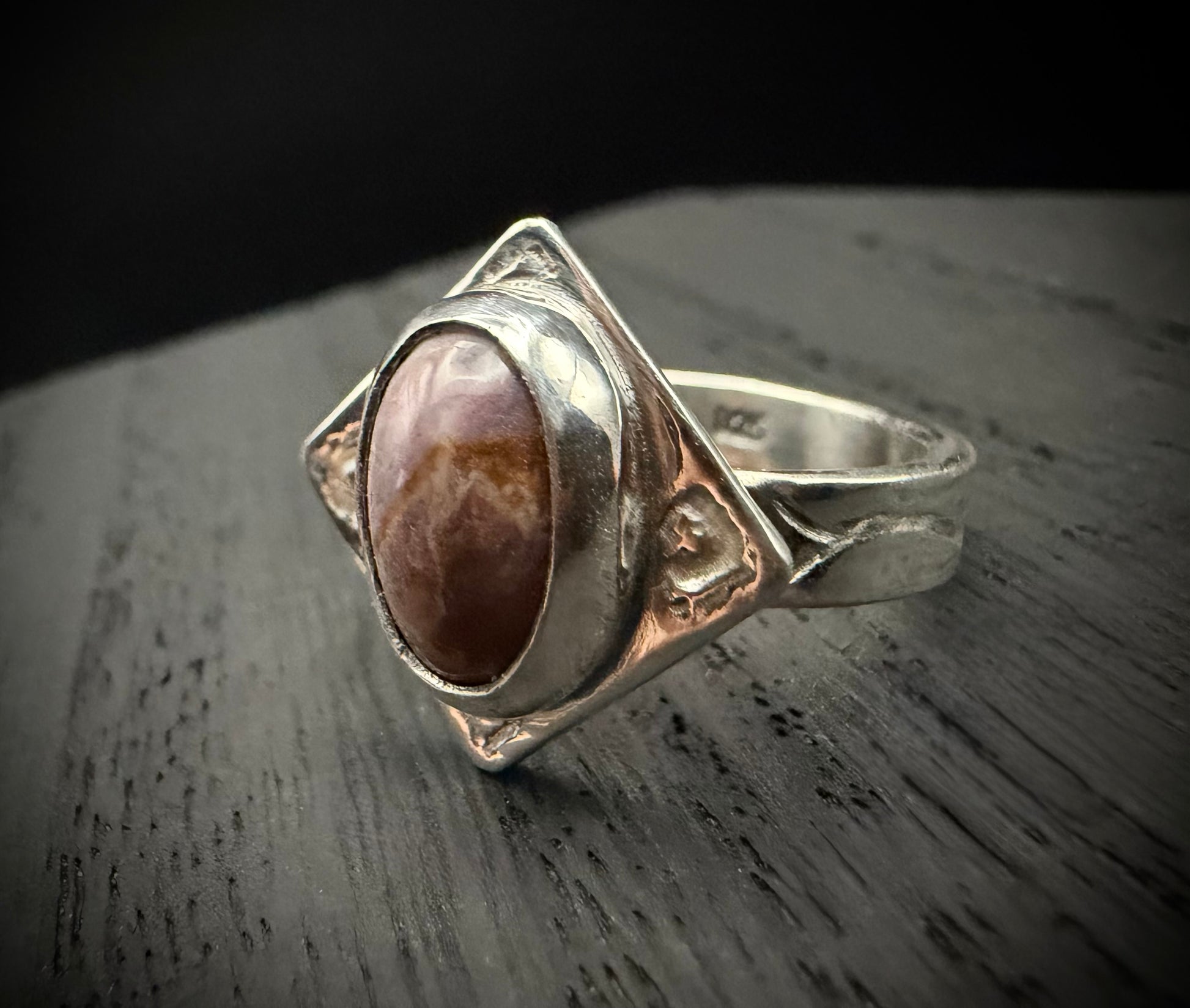 A close-up of the handcrafted sterling silver Tephra Ring resting on a dark, weathered wood surface. The ring features a diamond-shaped plate with molten textures and an earth-toned agate stone, highlighting the contrast between the polished silver edges and the deep brown of the gemstone.