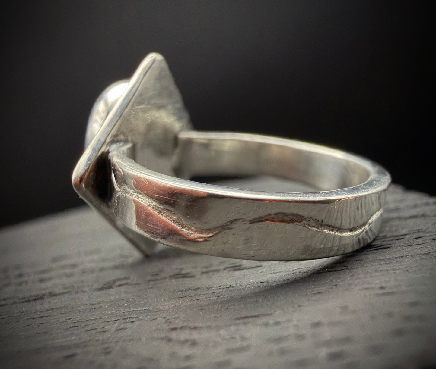 A profile view of the handcrafted sterling silver Tephra Ring resting on a dark, textured surface. The image highlights the solid silver band with its deep, undulating line that mimics the irregular flow of liquid metal.