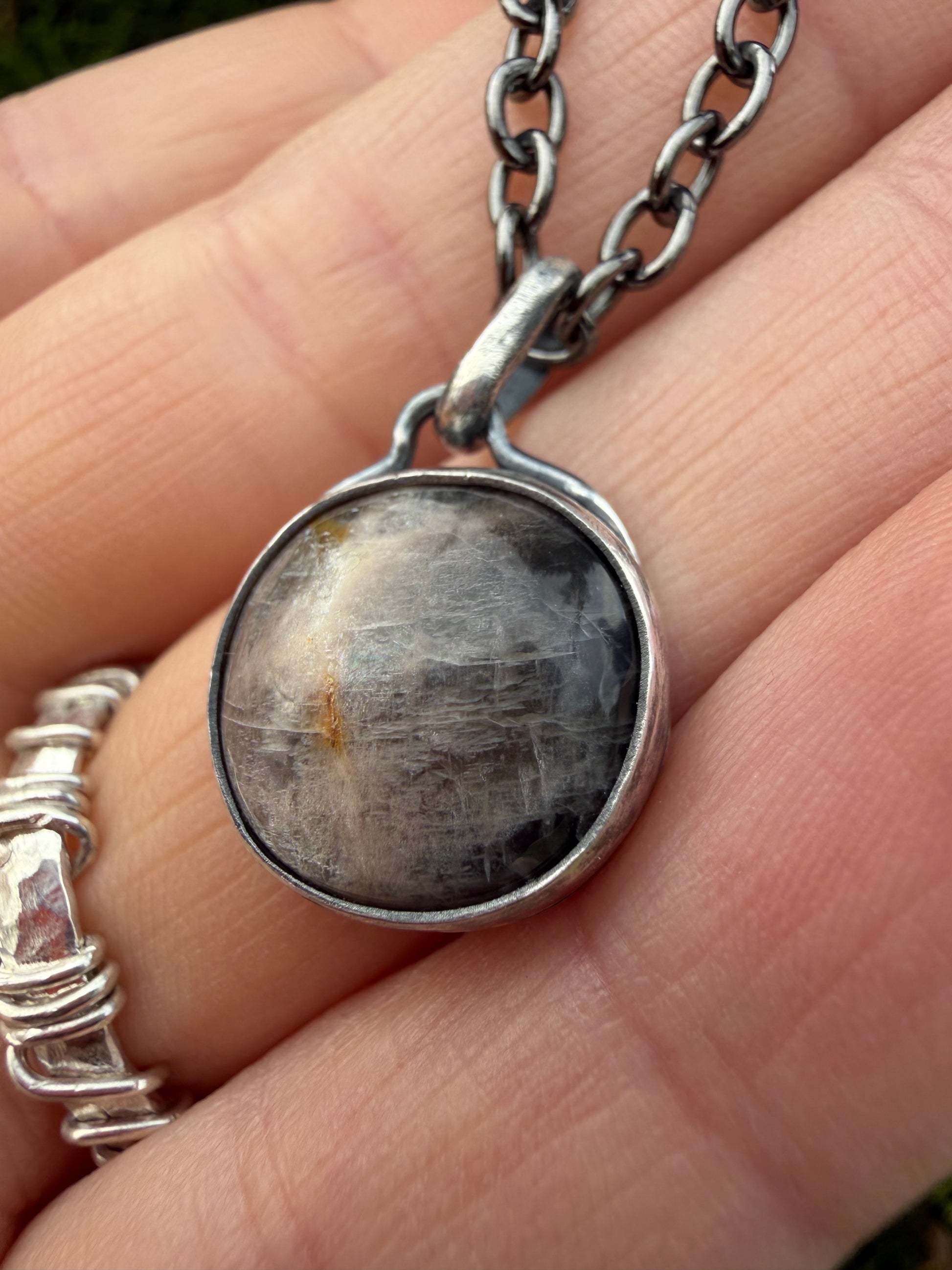 A close-up shot of the handcrafted sterling silver Lucian pendant held between fingers, with a partial view of a wrapped silver ring on the pinky finger. The round black moonstone features subtle, light-catching internal inclusions and a faint golden flash on the left side, all secured within a smooth bezel setting. A darkened patina silver chain attaches directly to a simple, classic loop bale at the top.