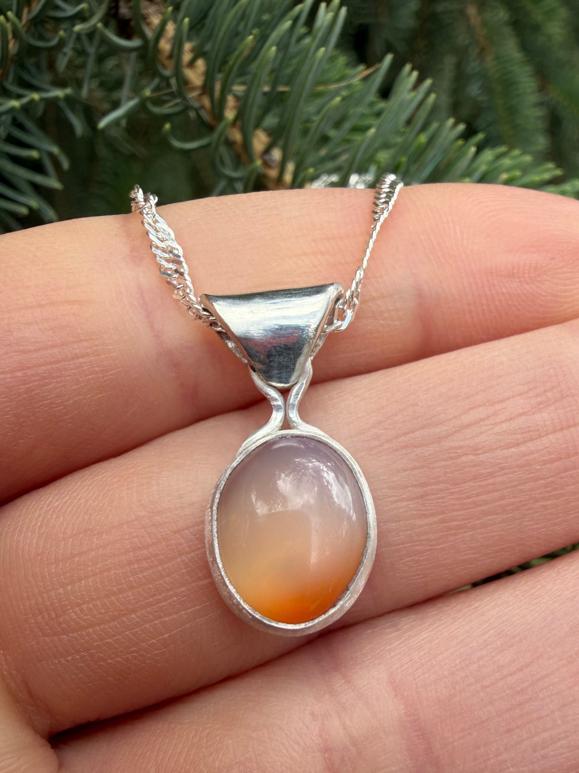 A close-up of the handcrafted sterling silver Lavinia pendant held between fingers against a soft background of evergreen pine needles. The polished, translucent banded carnelian chalcedony stone is secured in a smooth bezel setting, showcasing its rich orange tones and clear, parallel white banding.