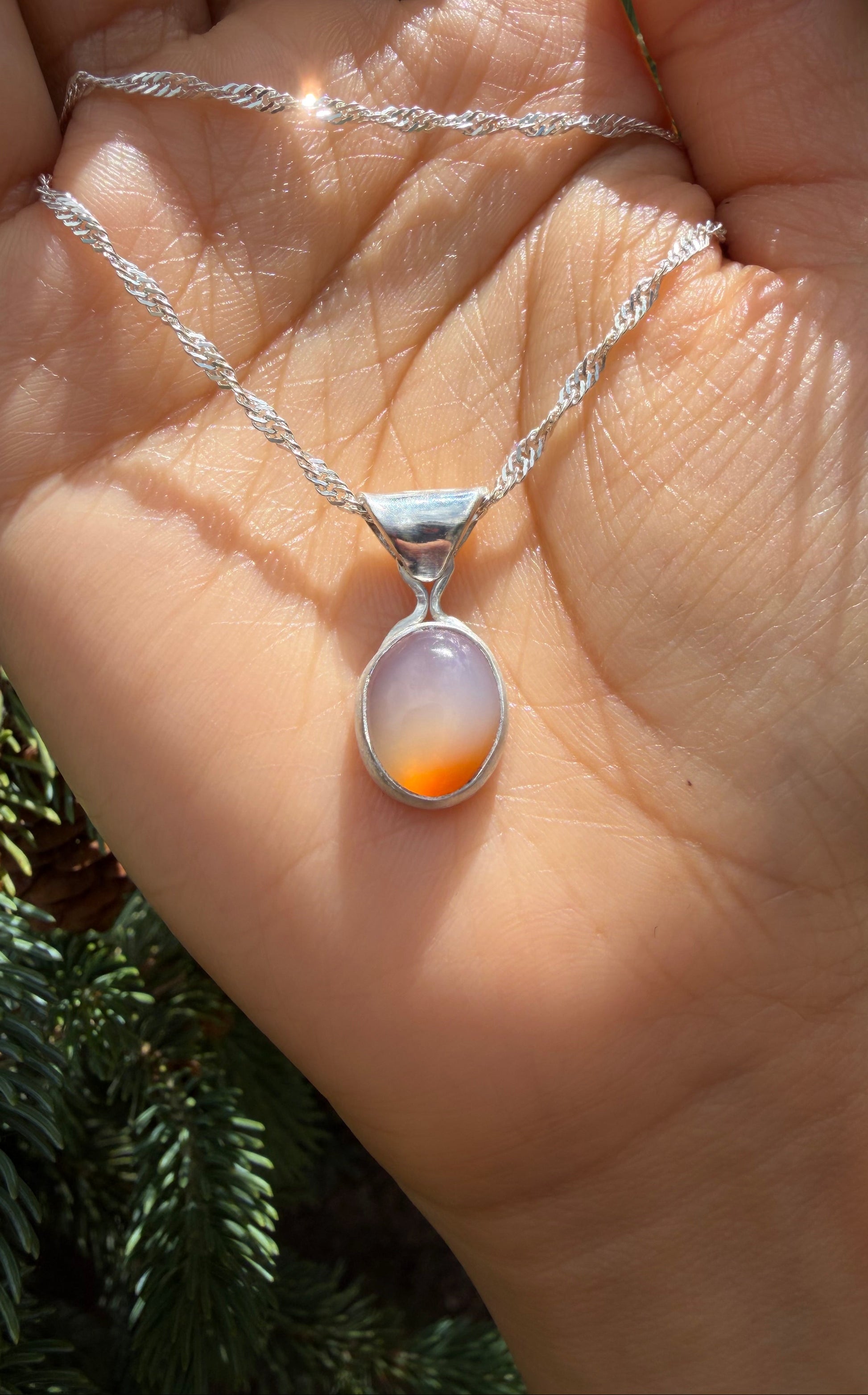 A close-up of the handcrafted sterling silver Lavinia pendant resting in an open palm. The dainty, sparkling silver chain is draped loosely across the hand, drawing focus to the polished oval setting. The banded carnelian chalcedony stone features a translucent, milky-white top half that transitions into a vibrant orange base. A soft background of evergreen pine needles is visible to the left.
