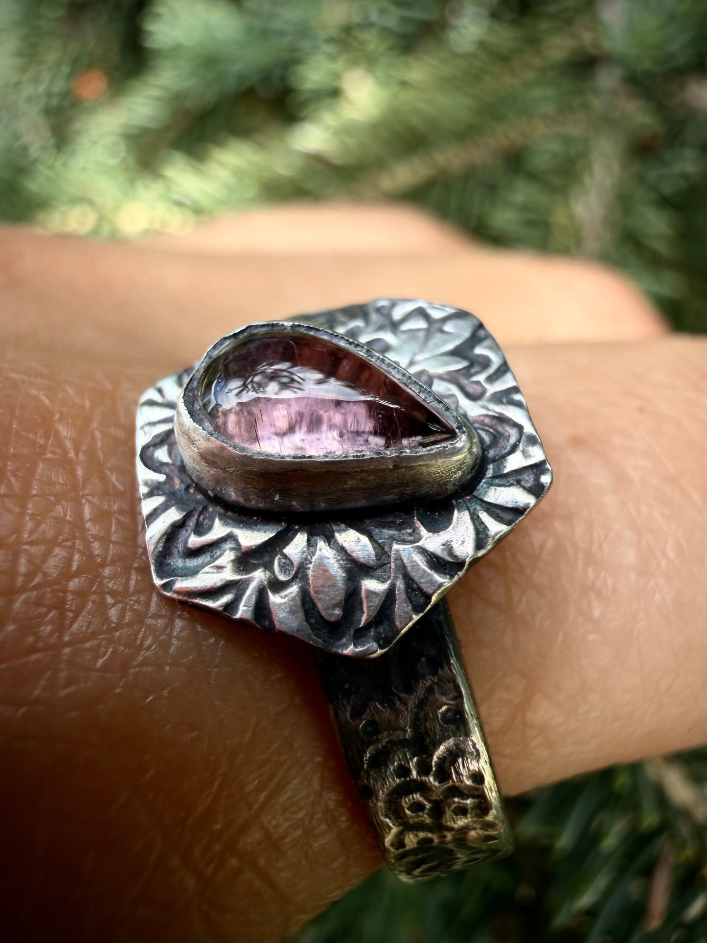 A close-up of the handcrafted sterling silver Nessa ring worn on a finger against a blurred green evergreen background. The ring features a horizontal, teardrop-shaped pink tourmaline stone secured in a fine silver bezel. The stone is framed by an ornate, textured silver border with a deep, darkened patina that highlights the detailed, organic patterns.