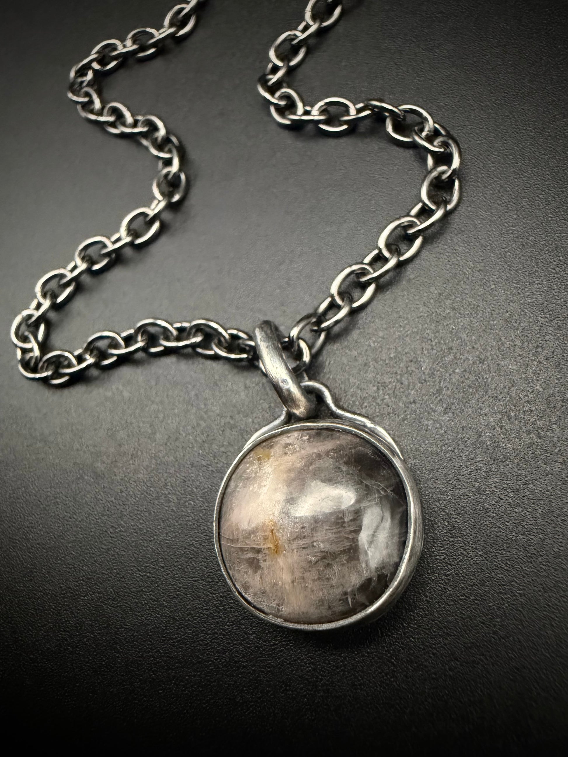 A close-up of the handcrafted sterling silver Lucian pendant resting on a dark, textured background. The round black moonstone is secured in a fine silver bezel, showcasing its dark, luminous tones and light-catching inclusions. The surrounding sterling silver features a deep patina finish, with the silver chain elegantly curved above the bale.