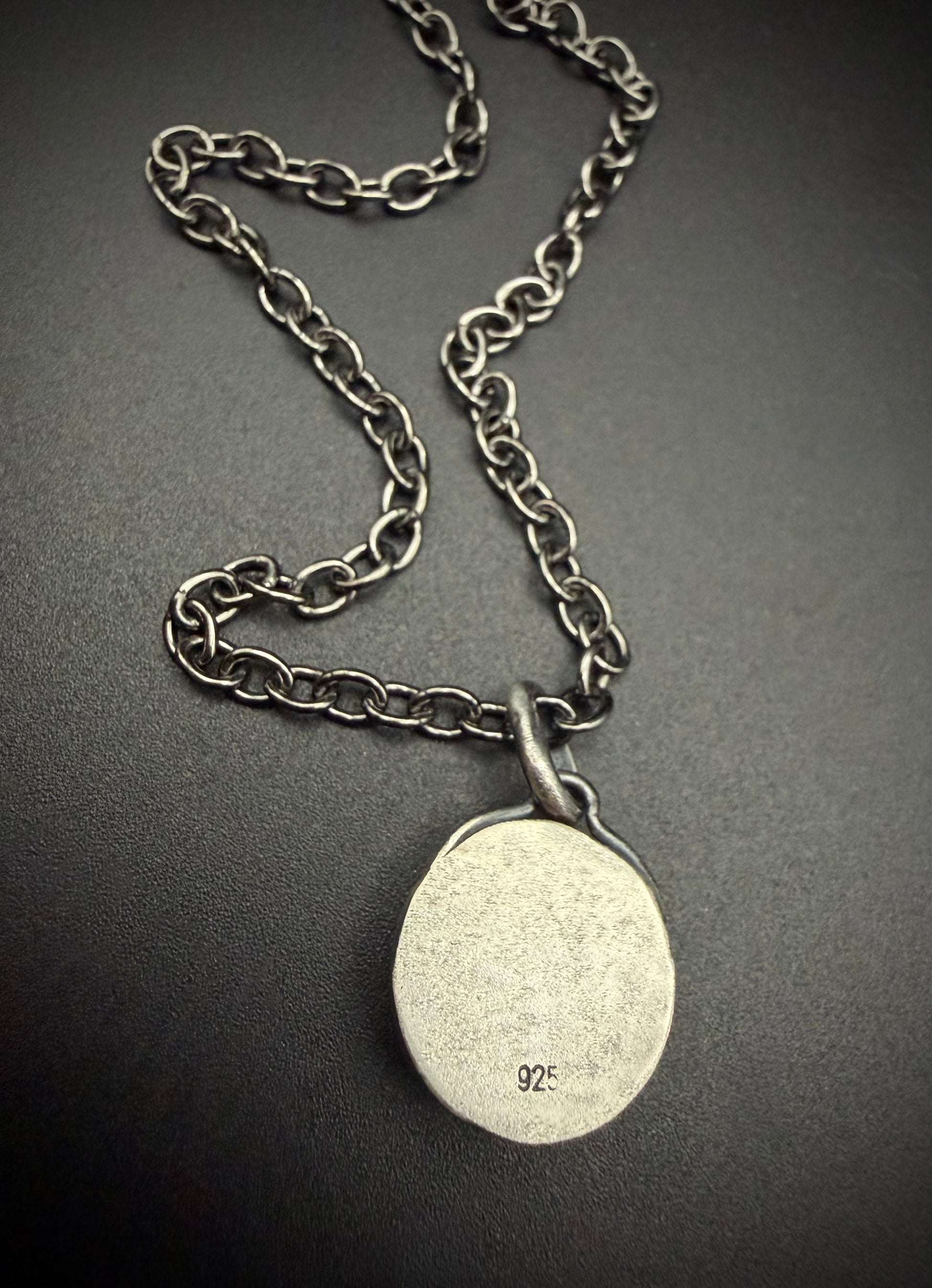 The reverse side of the handcrafted sterling silver Lucian pendant against a dark, matte background. The image highlights the "925" hallmark stamped cleanly near the bottom of the smooth, round silver backplate. The darkened patina silver link chain is elegantly curved above the simple, classic loop bale, showcasing the solid construction of the piece.