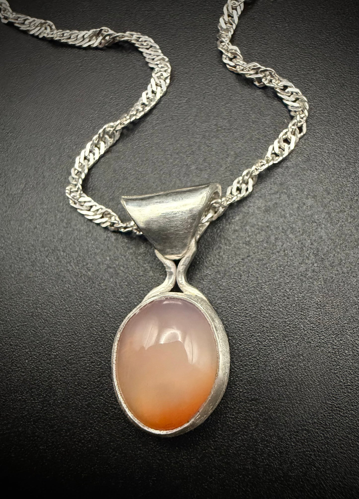 A close-up of the handcrafted sterling silver Lavinia pendant resting on a dark, textured background. The polished silver setting features a flared bale and holds a smooth, oval banded carnelian chalcedony stone with translucent orange tones and distinct, parallel white banding. The silver chain is elegantly draped above the piece.