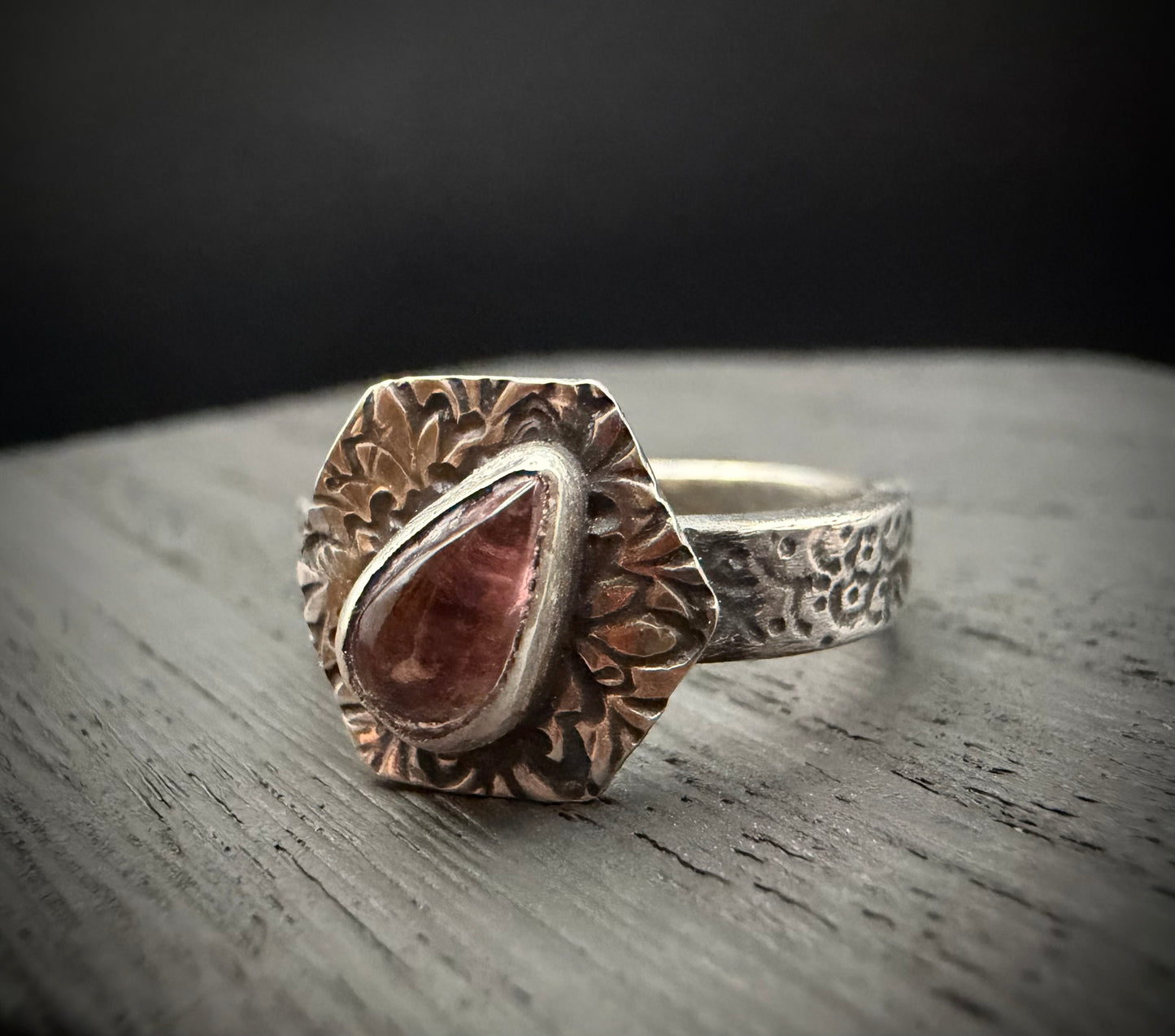 A close-up of the handcrafted sterling silver Nessa Ring resting on a textured, grey wooden surface. The ring features a teardrop-shaped pink tourmaline gemstone set inside a smooth bezel. The stone is centered on a prominent, hexagon-shaped face decorated with intricate, stylized floral patterns, attached to a textured, patterned silver band.