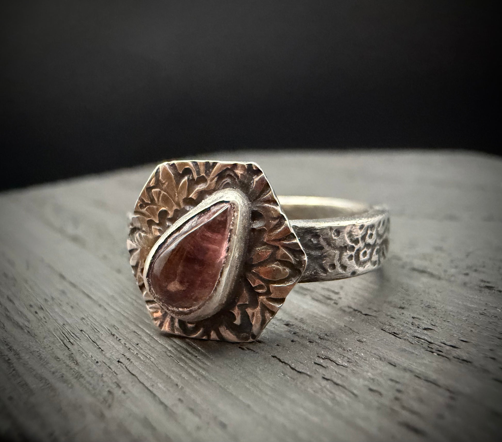 A close-up of the handcrafted sterling silver Nessa Ring resting on a textured, grey wooden surface. The ring features a teardrop-shaped pink tourmaline gemstone set inside a smooth bezel. The stone is centered on a prominent, hexagon-shaped face decorated with intricate, stylized floral patterns, attached to a textured, patterned silver band.