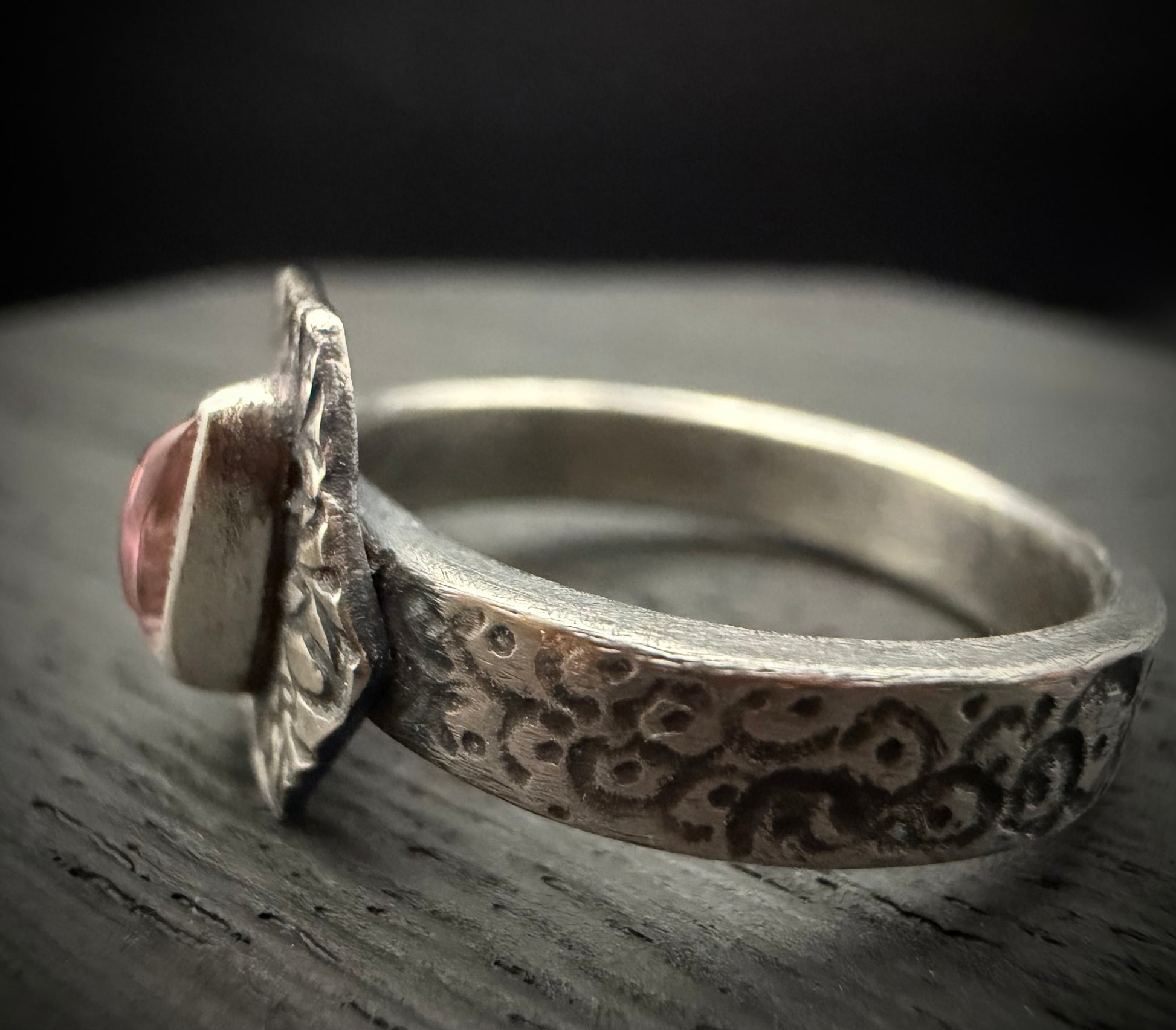 A side-profile view of the handcrafted sterling silver Nessa Ring resting on a dark, weathered wooden surface. The shot focuses on the thick ring band, which features a heavily textured, patterned surface with a dark patina in the recesses. A side profile of the teardrop-shaped pink tourmaline gemstone is visible, secured within its smooth silver bezel on the raised, decorative face.