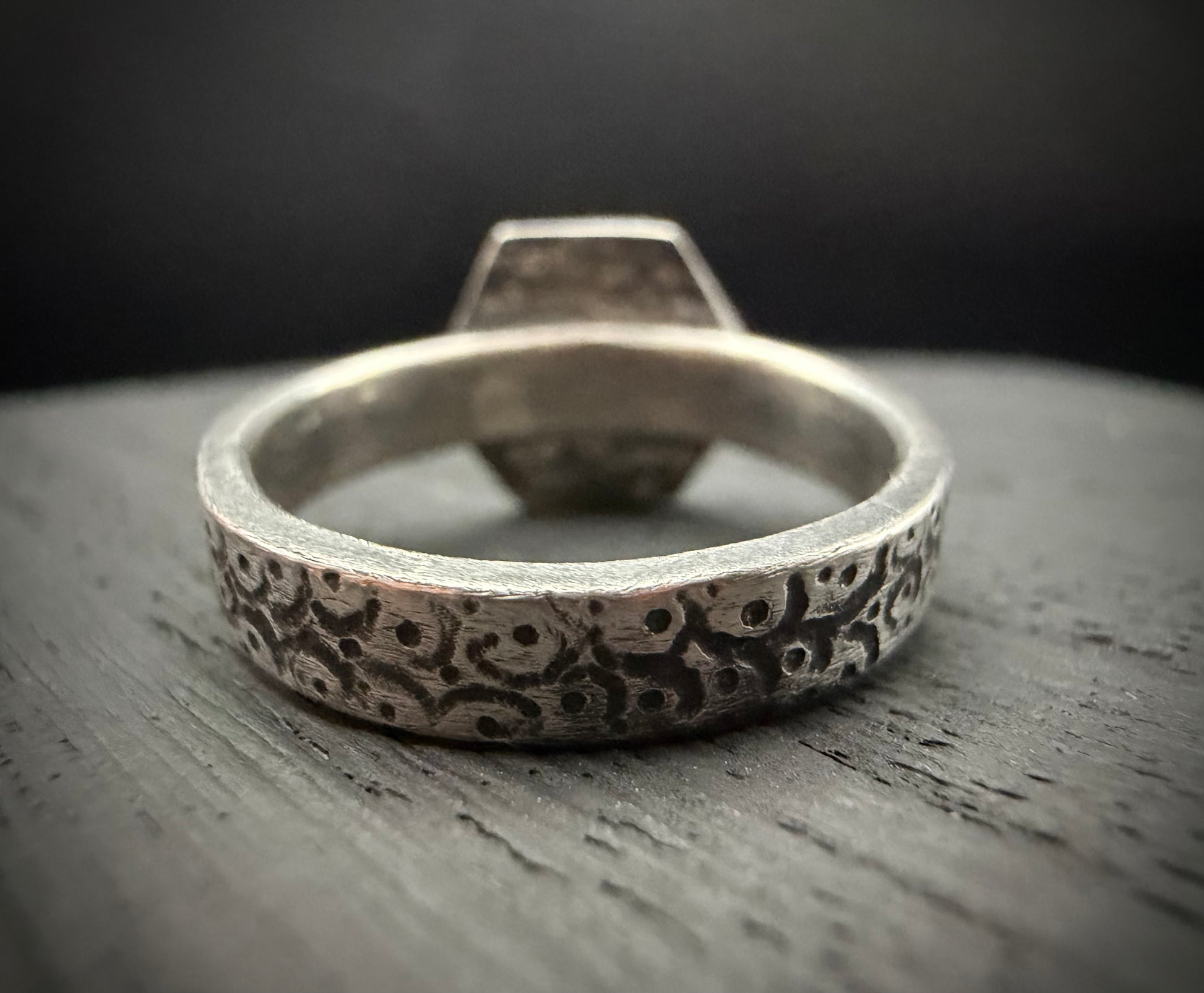A macro close-up view focusing on the back of the handcrafted sterling silver Nessa Ring band resting on a textured, grey surface. The shot highlights the thick, rounded profile of the band, featuring an intricate, deeply recessed pattern with a dark patina finish. The raised hexagon-shaped face of the setting rises up in the background, softly out of focus.