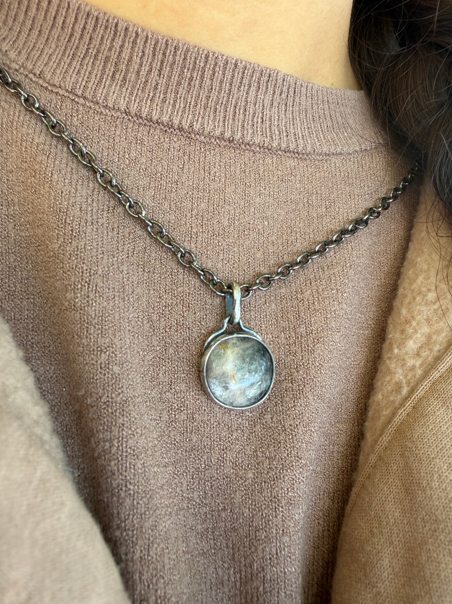 A close-up lifestyle shot of the handcrafted sterling silver Lucian pendant being worn against a beige knit sweater. The round black moonstone is secured in a smooth silver bezel, showcasing its dark, luminous tones and light-catching inclusions. The surrounding sterling silver and matching chain feature a deep patina finish, with the chain looping cleanly through a classic, simple bale.