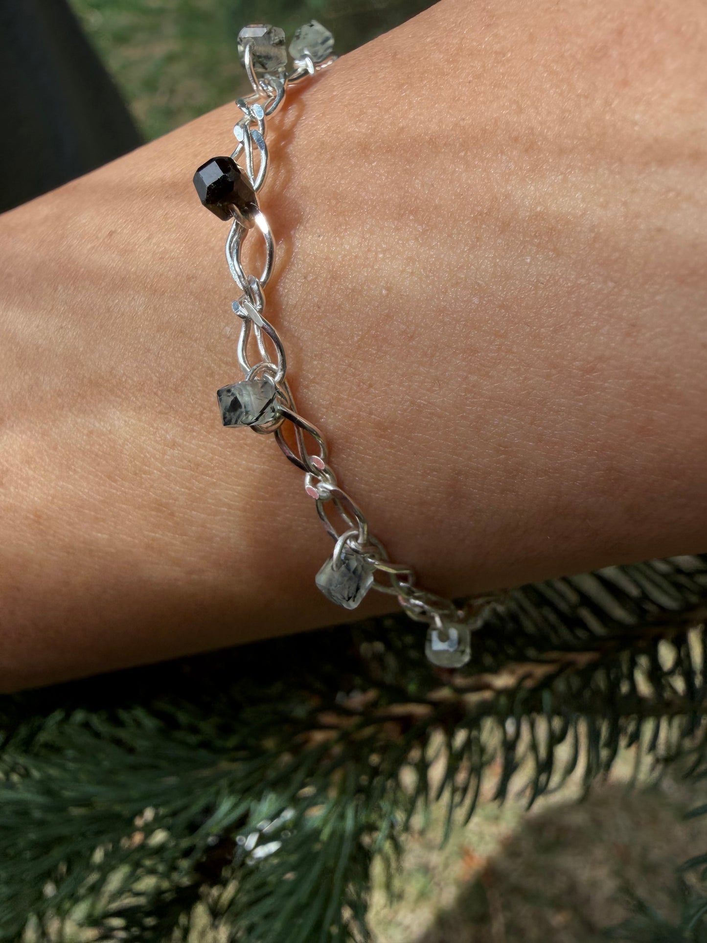 A close-up photograph of the handcrafted Lumos Bracelet worn on a wrist, positioned against a background of natural green pine needles. The bright, shiny sterling silver chain catches the light, showcasing translucent, ice-like prehnite stone beads threaded onto the links—including a prominent dark, faceted inclusion-rich gemstone bead.
