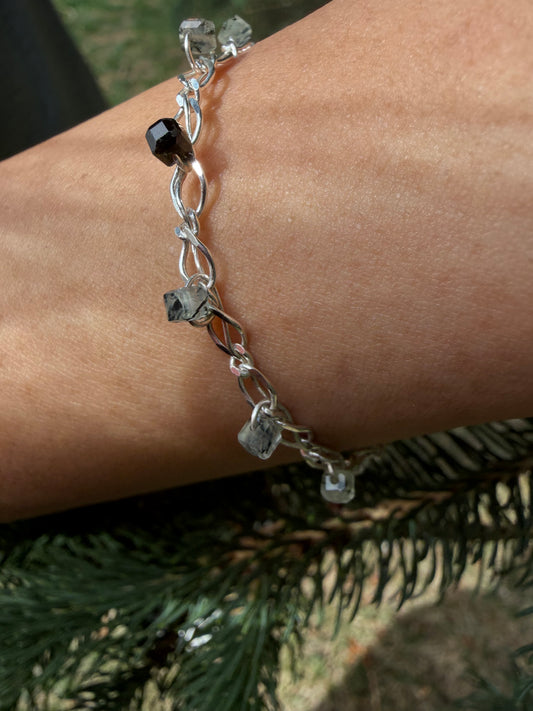 A close-up photograph of the handcrafted Lumos Bracelet worn on a wrist, positioned against a background of natural green pine needles. The bright, shiny sterling silver chain catches the light, showcasing translucent, ice-like prehnite stone beads threaded onto the links—including a prominent dark, faceted inclusion-rich gemstone bead.