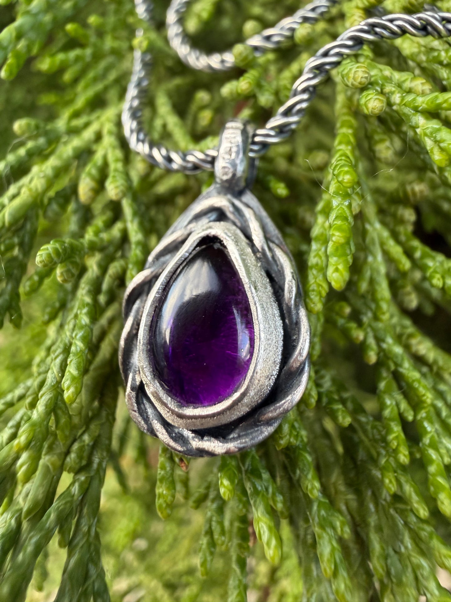 A close-up photograph of the handcrafted sterling silver Nodus Pendant worn on a model. The teardrop-shaped pendant features a vibrant, polished purple amethyst gemstone nestled inside a heavy, twisted silver rope frame, with a dark patina highlighting the deep grooves of the metalwork.