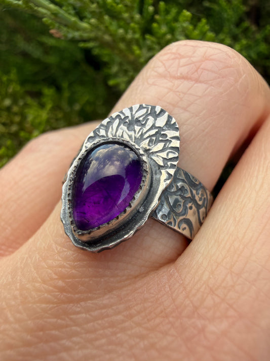A close-up of the handcrafted sterling silver Nightshade Ring being worn on a finger, set against a soft, blurred green evergreen background. The ring features a large, teardrop-shaped purple amethyst gemstone secured in a fine silver bezel. The stone is topped by an intricate, fan-shaped silver backplate featuring a detailed, stylized tree or branching plant pattern with a dark patina that highlights the fine metalwork. The wide ring band is also heavily textured with a deeply recessed, organic swirling pa