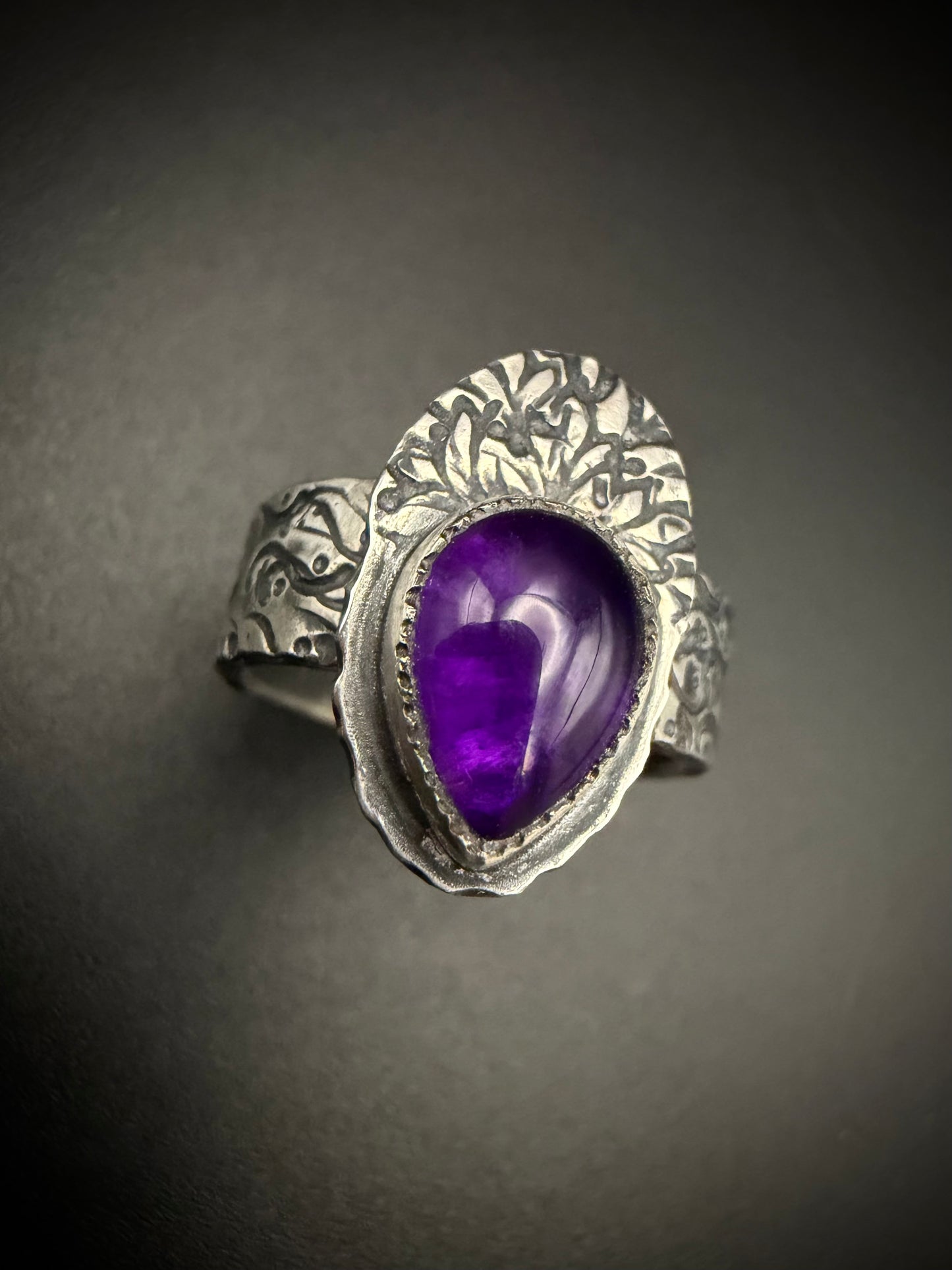 A close-up view of the handcrafted sterling silver Nightshade Ring resting on a dark gray surface. The shot centers on the large, teardrop-shaped purple amethyst gemstone secured in its smooth fine silver bezel, highlighting the prominent, fan-shaped backplate decorated with an intricate plant or tree branch pattern.