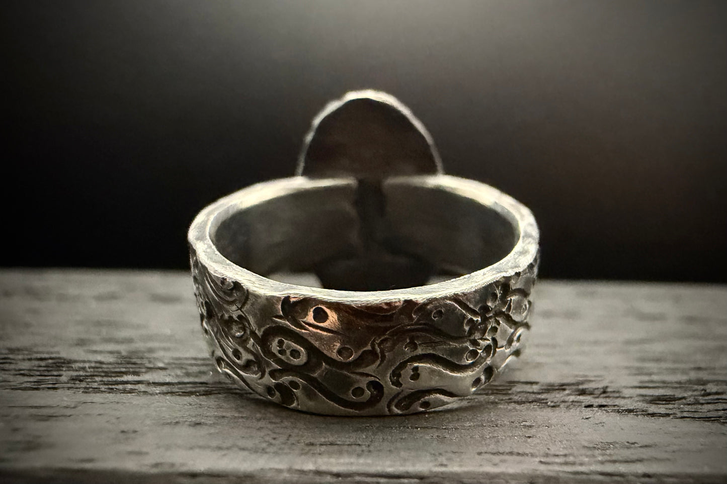 A macro close-up view of the back of the handcrafted sterling silver Nightshade Ring band, resting on a rustic, weathered wooden surface. The shot highlights the wide, thick profile of the band, showcasing a deeply recessed, organic swirling pattern with a dark patina finish. The plain, unpatinated silver back of the setting rises up in the background, softly out of focus.