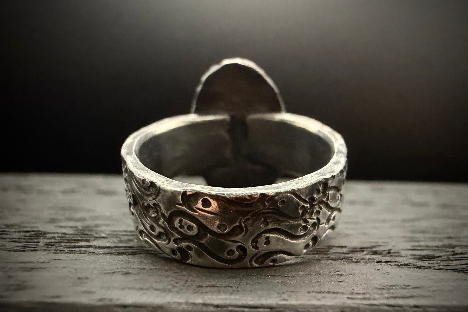 A macro close-up view of the back of the handcrafted sterling silver Nightshade Ring band, resting on a rustic, weathered wooden surface. The shot highlights the wide, thick profile of the band, showcasing a deeply recessed, organic swirling pattern with a dark patina finish. The plain, unpatinated silver back of the setting rises up in the background, softly out of focus.