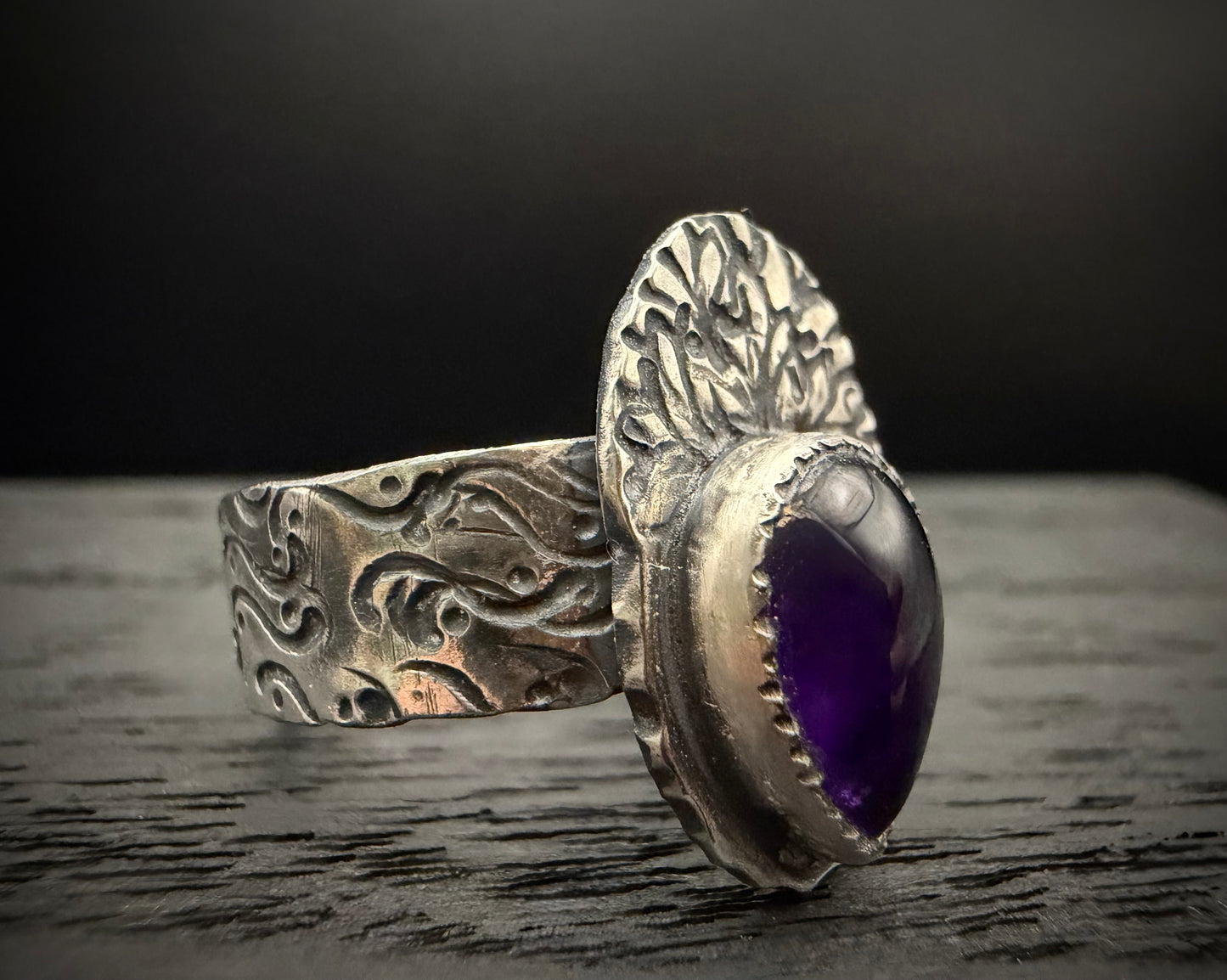 A top-down view of the handcrafted sterling silver Nightshade Ring resting flat on a dark surface. The shot clearly showcases the unique symmetry of the fan-shaped backplate and its intricate branching plant pattern, contrasting against the rich purple of the teardrop amethyst gemstone.