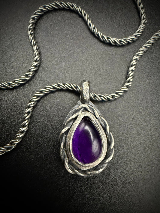 A close-up photograph of the handcrafted sterling silver Nodus Pendant resting on a dark, textured surface alongside its matching twisted rope chain. The teardrop-shaped pendant features a vibrant, polished purple amethyst gemstone nestled inside a heavy frame of intricately coiled and twisted silver wire, with a dark patina highlighting the deep grooves of the metalwork.