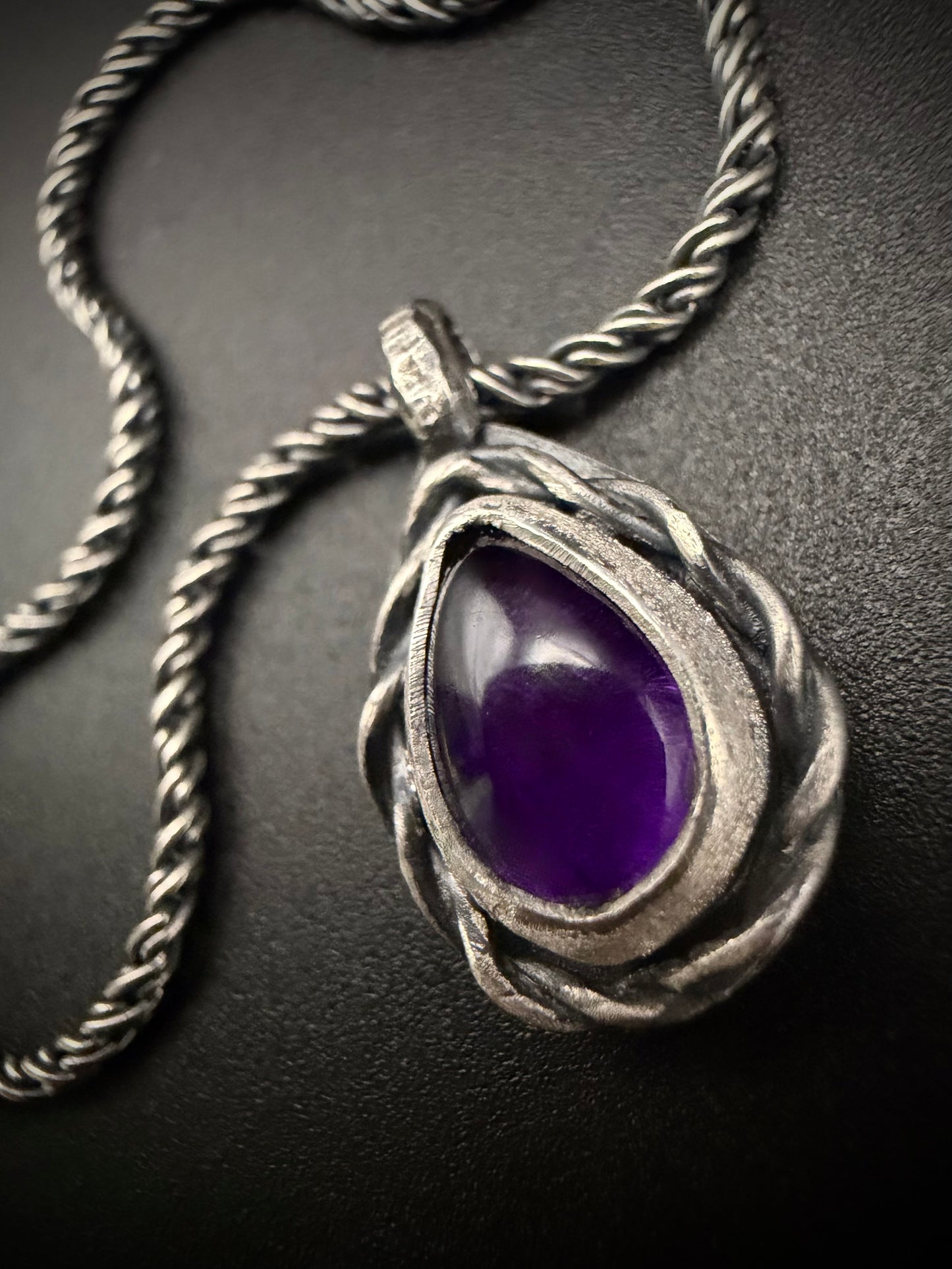 A close-up photograph of the handcrafted sterling silver Nodus Pendant. The teardrop-shaped pendant features a vibrant, polished purple amethyst gemstone nestled inside a heavy, twisted silver rope frame, with a dark patina highlighting the deep, textured grooves of the metalwork.