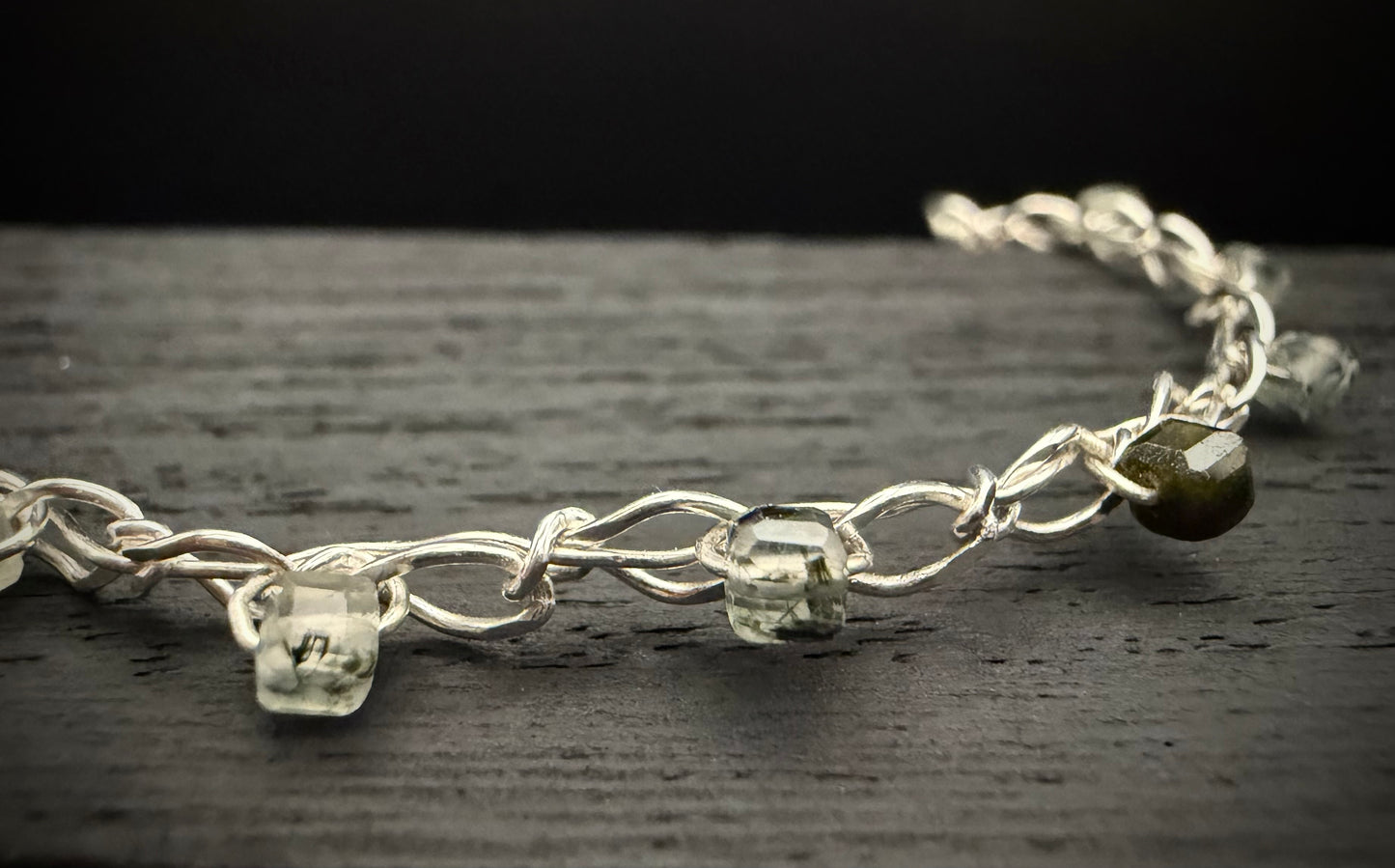 A macro close-up of a segment of the bright, shiny sterling silver chain from the handcrafted Lumos Bracelet resting on a dark, textured surface. The focus centers on two clear, translucent pale green prehnite cubes and one darker, olive-green inclusion-rich prehnite bead threaded onto the intricately linked chain.