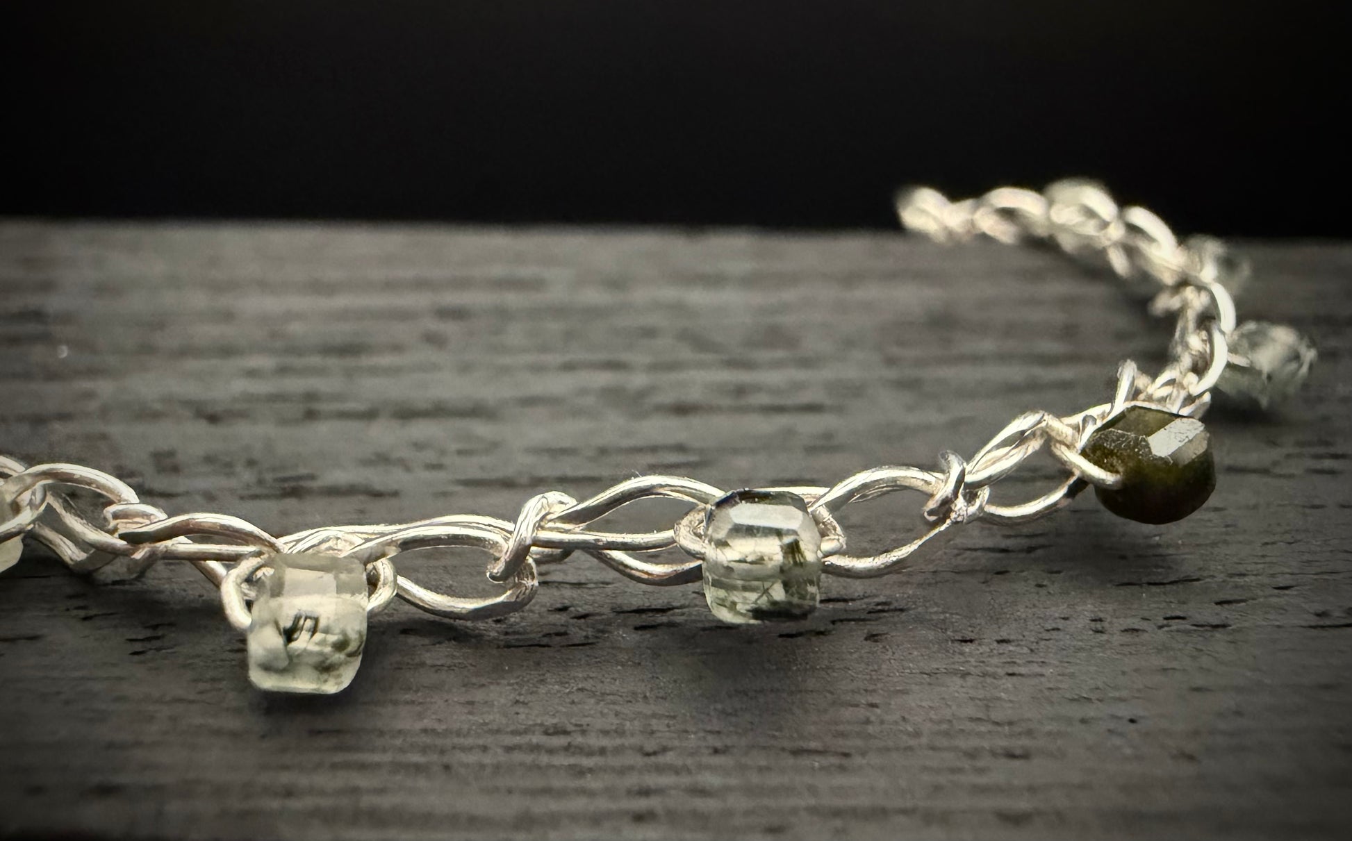 A macro close-up of a segment of the bright, shiny sterling silver chain from the handcrafted Lumos Bracelet resting on a dark, textured surface. The focus centers on two clear, translucent pale green prehnite cubes and one darker, olive-green inclusion-rich prehnite bead threaded onto the intricately linked chain.