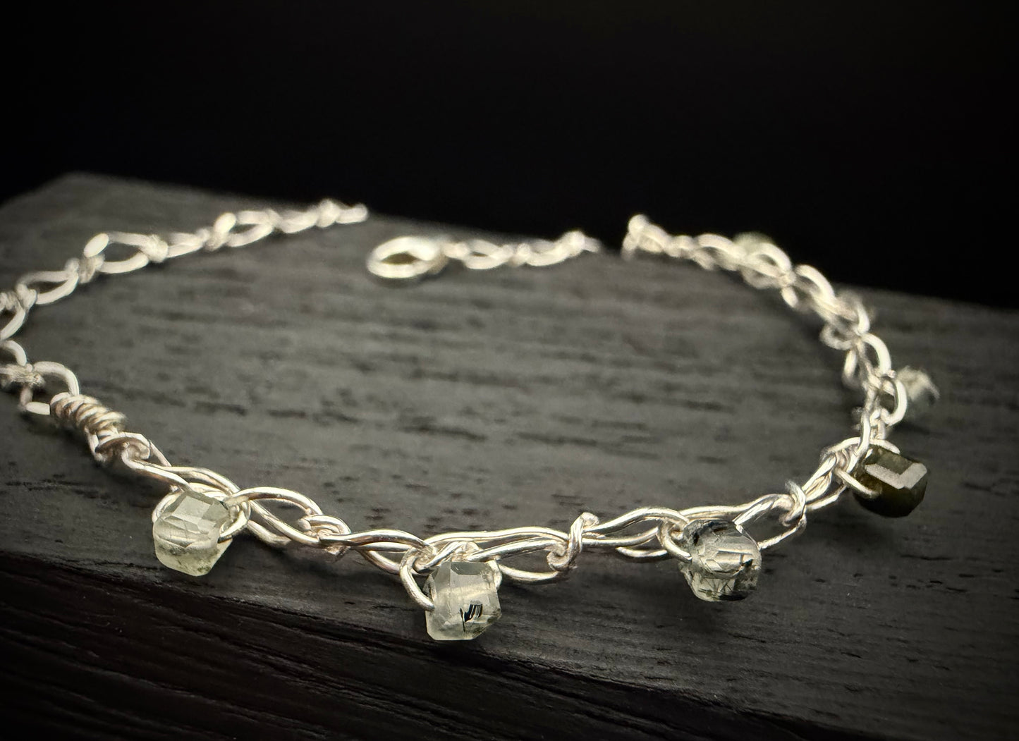 A close-up view of the handcrafted sterling silver Lumos Bracelet resting on a dark, rustic wooden surface. The shot displays a segment of the bright, shiny classic sterling chain, featuring a few translucent, pale green prehnite beads that catch the light against the dark background.