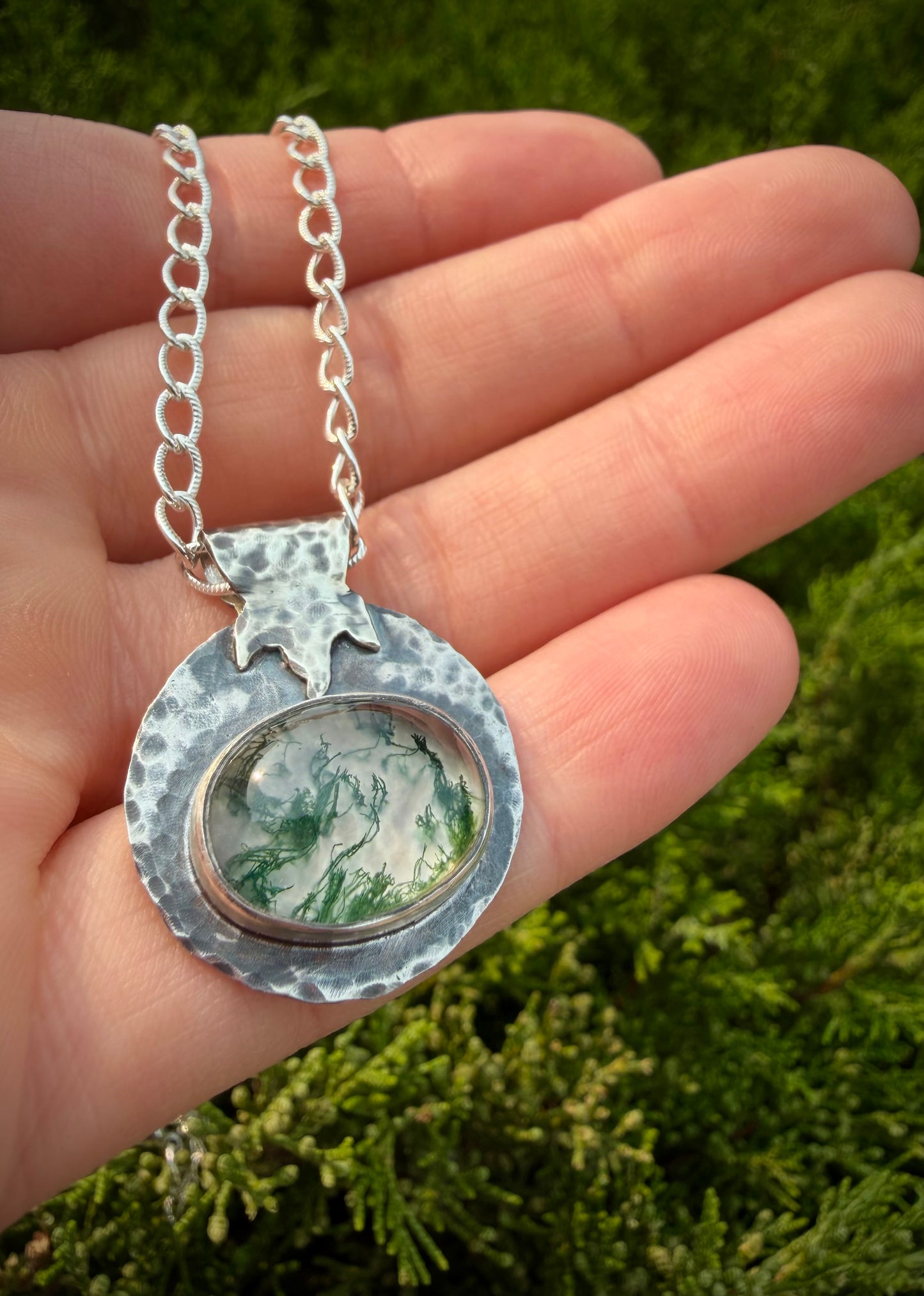Close-up of a handcrafted sterling silver pendant featuring a large, oval moss agate stone with intricate green, moss-like inclusions. The stone is set in a fine silver bezel, surrounded by a wide, heavily hammered, and textured silver border with a stylized leaf or flame bail, hanging from a thick silver chain.