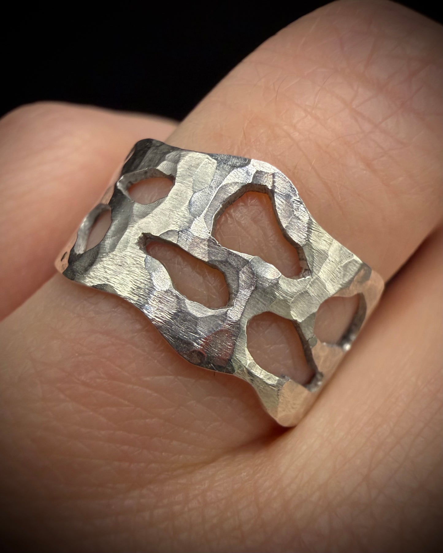 Close-up of a wide, handcrafted sterling silver ring worn on a finger. The ring features a heavily hammered, textured surface with unique, organic-shaped geometric cutouts showcasing the skin beneath.
