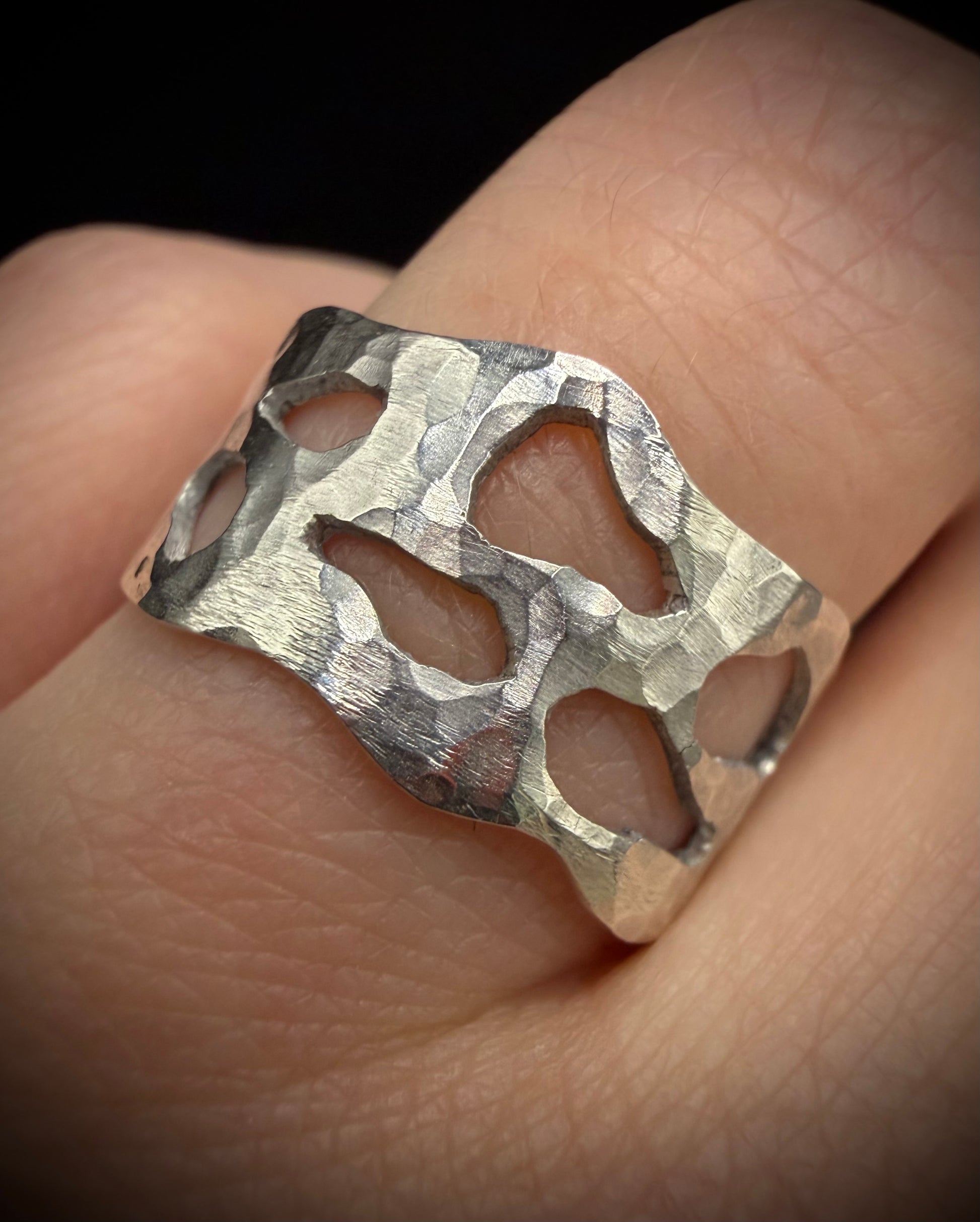 Close-up of a wide, handcrafted sterling silver ring worn on a finger. The ring features a heavily hammered, textured surface with unique, organic-shaped geometric cutouts showcasing the skin beneath.
