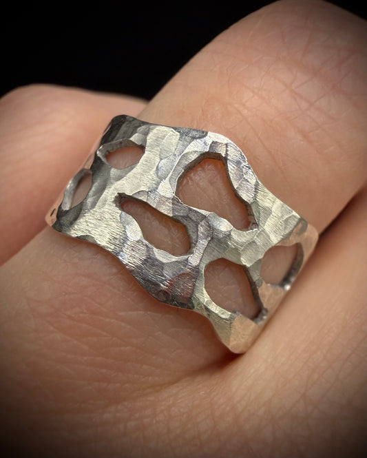 Close-up of a wide, handcrafted sterling silver ring worn on a finger. The ring features a heavily hammered, textured surface with unique, organic-shaped geometric cutouts showcasing the skin beneath.
