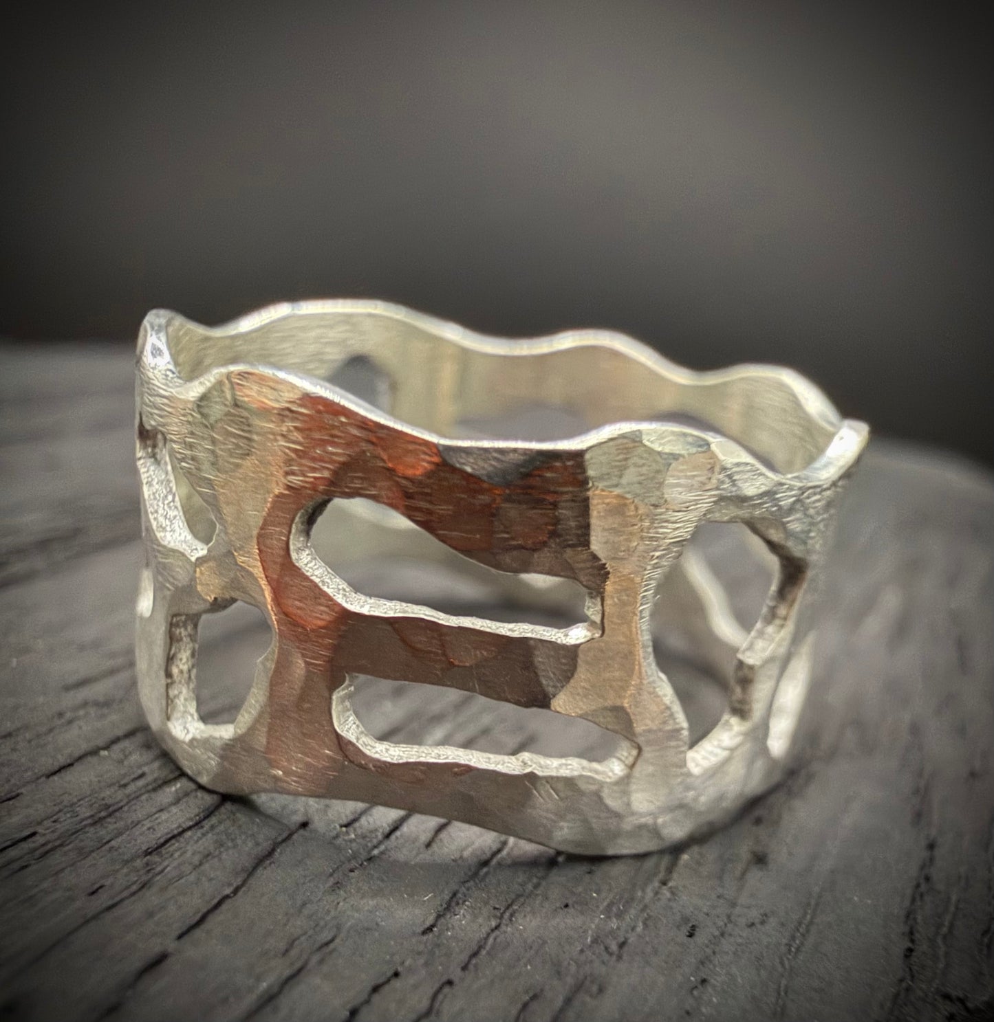 Top-down view of a wide, handcrafted sterling silver ring resting on a dark, rustic wooden surface. The ring features a heavily hammered, textured finish and bold geometric cutouts that reveal the wood grain underneath.
