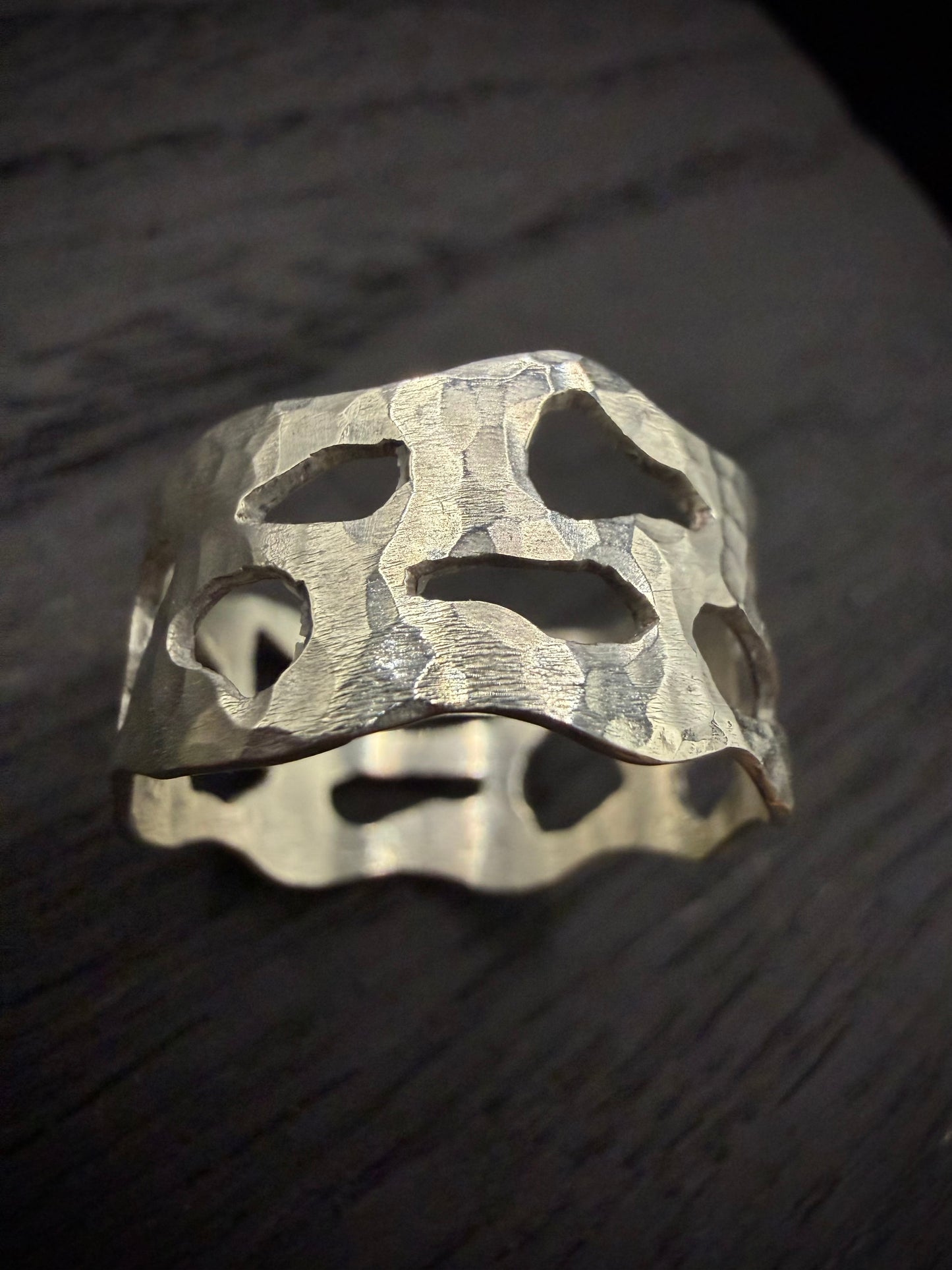 Studio shot of a wide, handcrafted sterling silver band ring displayed on a dark background. The ring showcases a distinctive, heavily hammered texture and broad, organic geometric cutouts that create an edgy, modern look.