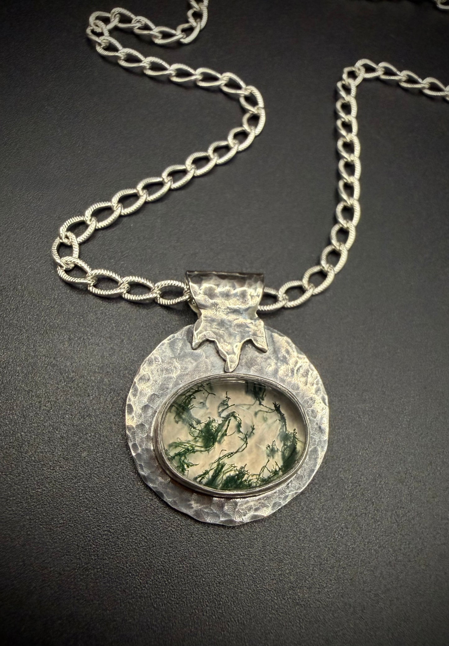 Close-up of a handcrafted sterling silver necklace featuring a round, heavily hammered pendant. The pendant showcases an oval moss agate gemstone with distinct green, branching moss-like patterns set inside a fine silver bezel, topped with a stylized, textured leaf bail on a thick silver chain.
