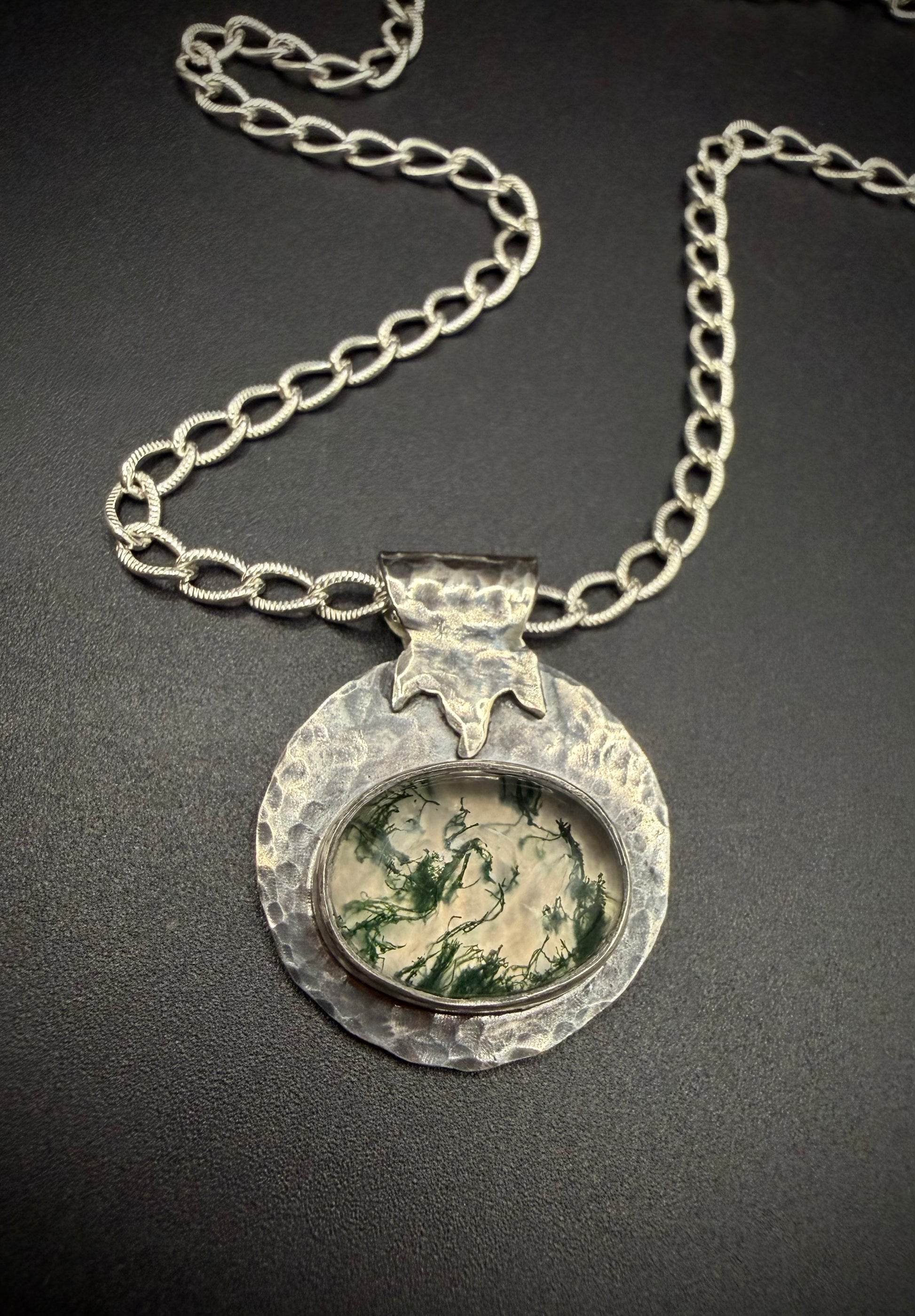 Close-up of a handcrafted sterling silver necklace featuring a round, heavily hammered pendant. The pendant showcases an oval moss agate gemstone with distinct green, branching moss-like patterns set inside a fine silver bezel, topped with a stylized, textured leaf bail on a thick silver chain.