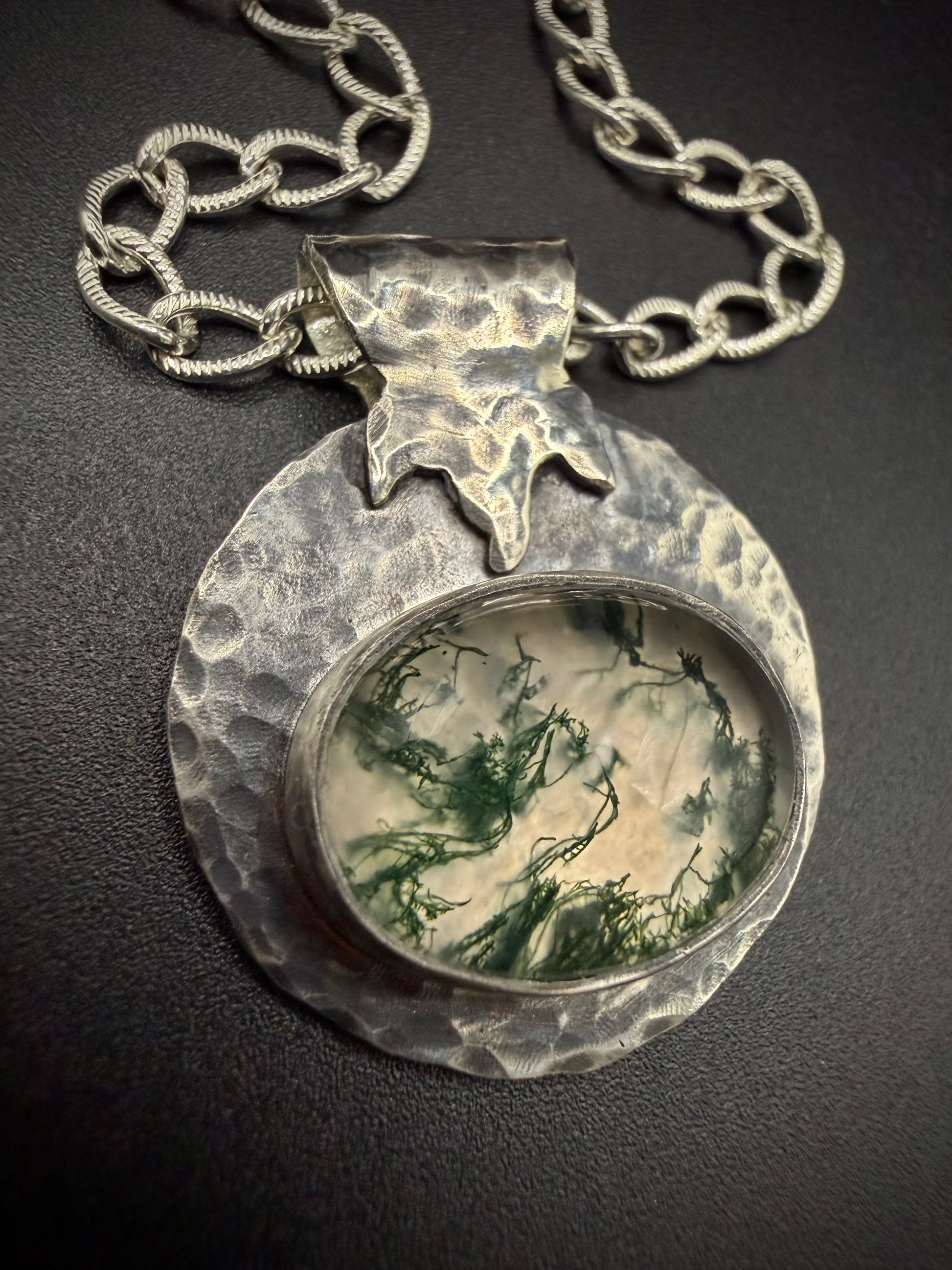 Close-up of a handcrafted sterling silver pendant featuring a large, oval moss agate stone with intricate green, moss-like inclusions. The stone is set in a fine silver bezel, surrounded by a wide, heavily hammered, and textured silver border with a stylized leaf or flame bail, hanging from a thick silver chain.