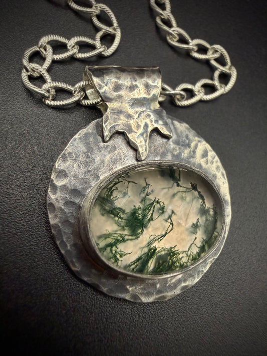 Close-up of a handcrafted sterling silver pendant featuring a large, oval moss agate stone with intricate green, moss-like inclusions. The stone is set in a fine silver bezel, surrounded by a wide, heavily hammered, and textured silver border with a stylized leaf or flame bail, hanging from a thick silver chain.