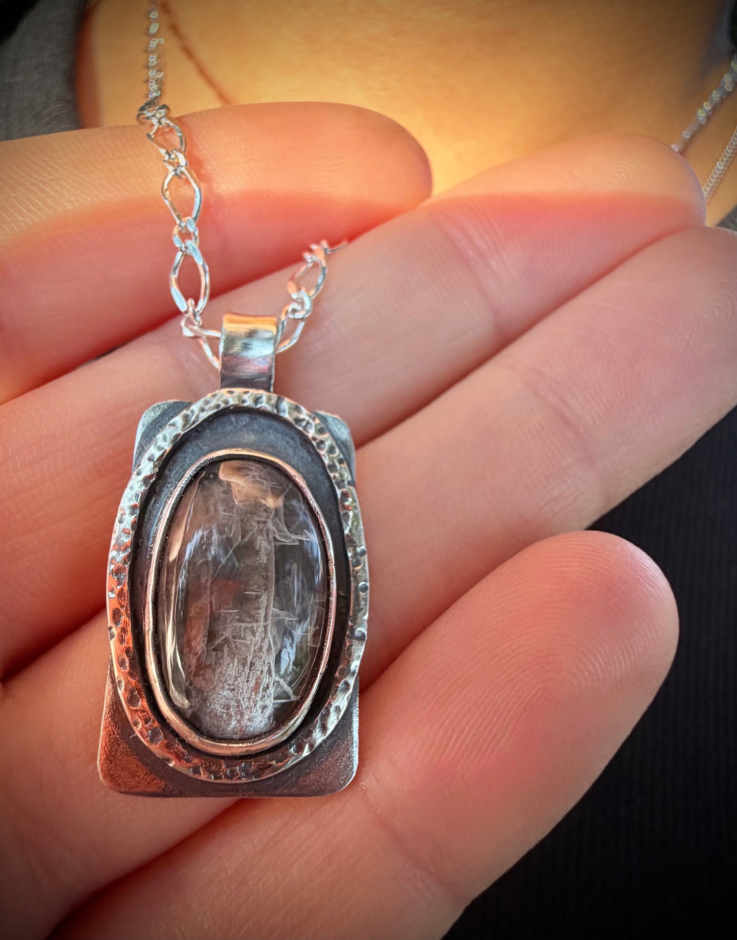 A close-up of a hand holding the Nova Pendant, a rectangular handcrafted sterling silver necklace featuring a prominent oval black moonstone cabochon set in a textured, multi-layered silver frame with a dark, moody patina.