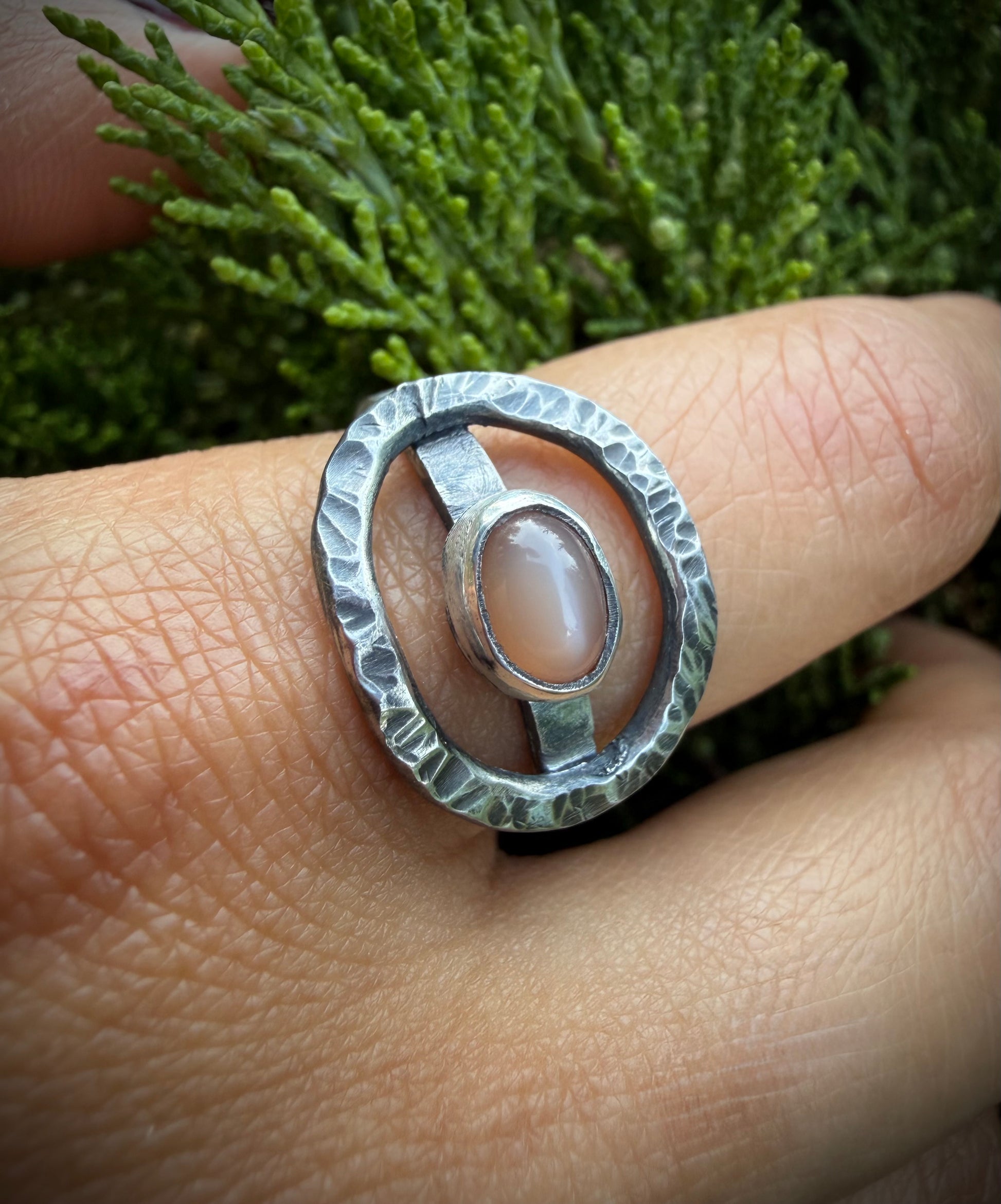 Close-up of a hand wearing a hammered sterling silver statement ring with an open oval design and a central peach moonstone stone, set against a green evergreen background.