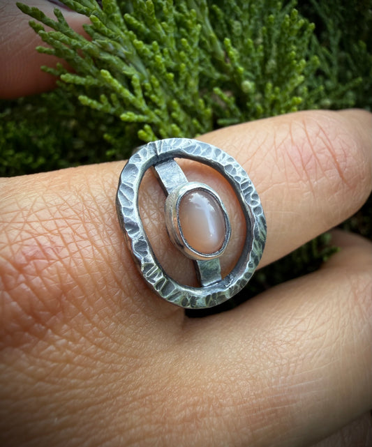 Close-up of a hand wearing a hammered sterling silver statement ring with an open oval design and a central peach moonstone stone, set against a green evergreen background.