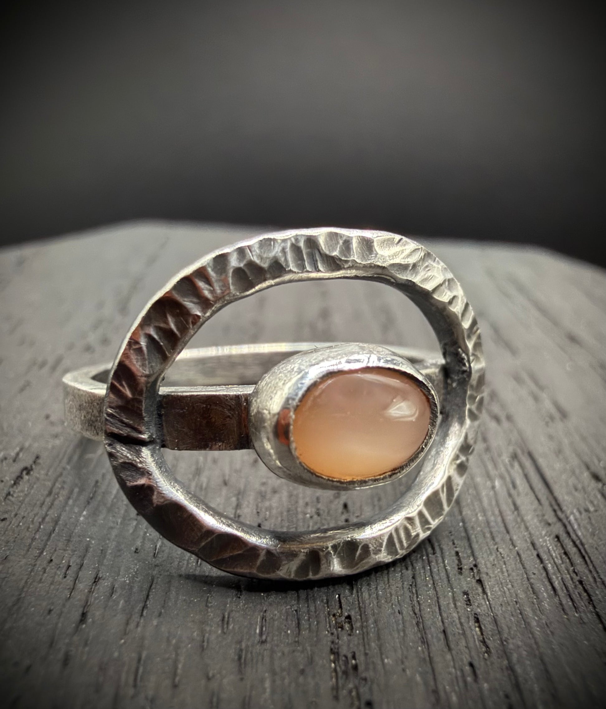 A close-up of a handcrafted sterling silver ring resting on a dark wood surface, featuring a textured, open circle frame with a weathered finish and a central oval peach moonstone cabochon set in a fine silver bezel.