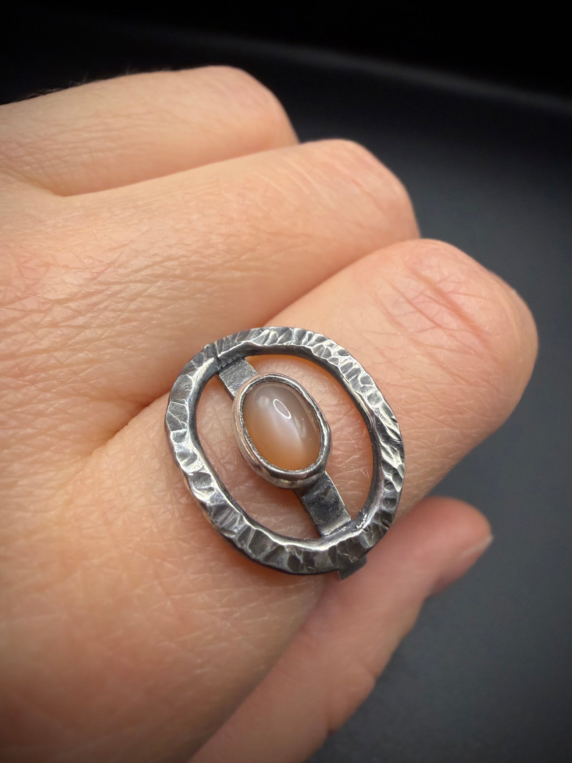 Close-up of a hand wearing the Cora Ring, a statement ring featuring an open, textured oval sterling silver frame with a weathered finish and a central peach moonstone cabochon set in a fine silver bezel.