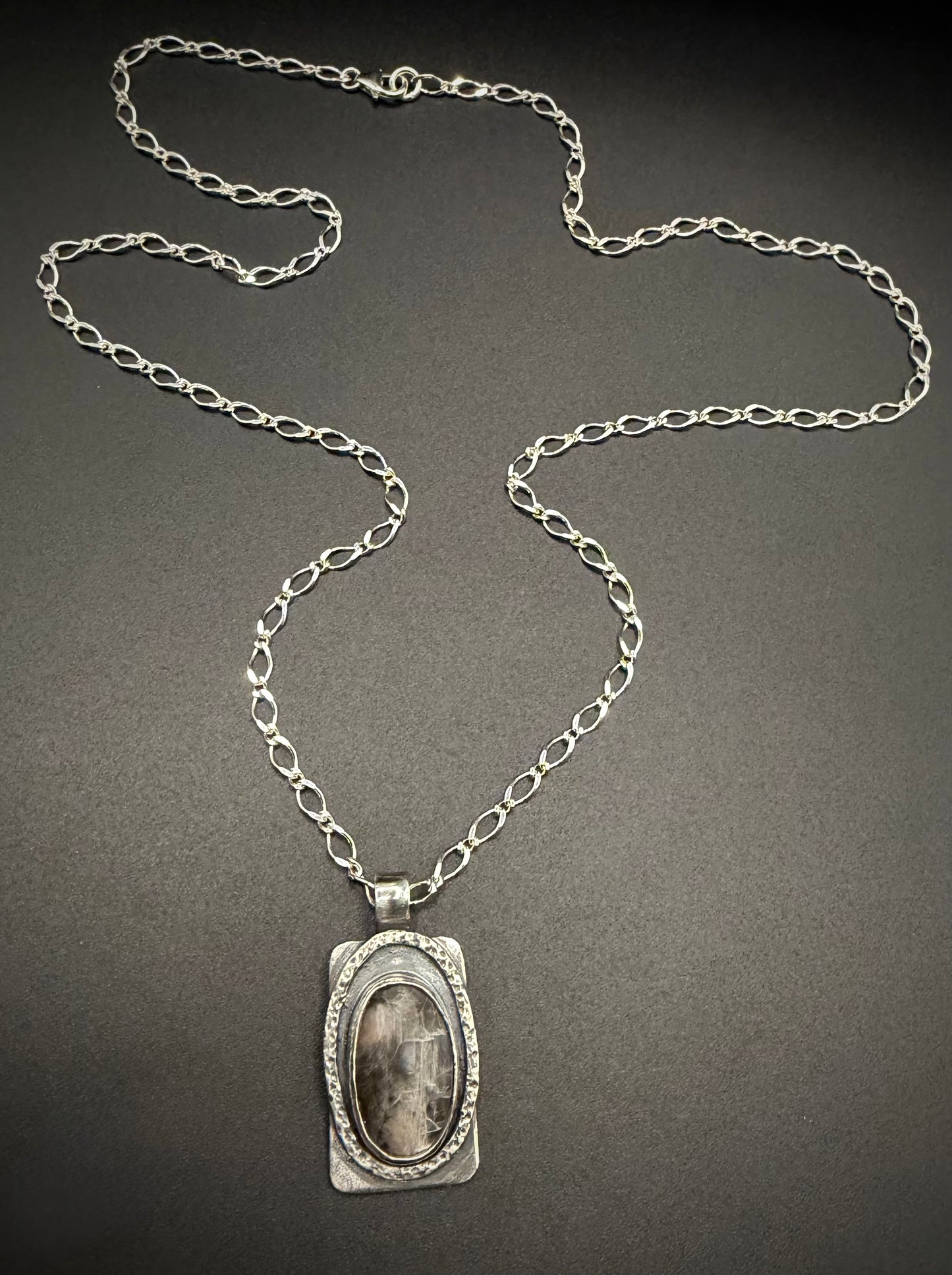 A handcrafted rectangular sterling silver pendant on a long link chain, resting against a dark background. The pendant features a central, vertically oriented oval black moonstone cabochon enclosed by a deeply textured, layered silver bezel.